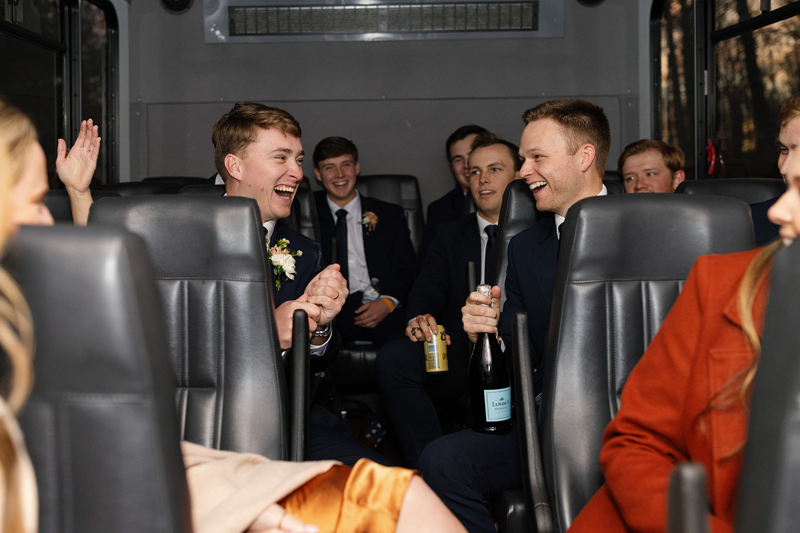 Groom laughs with groomsmen aboard the party bus, champagne and beers in hand.