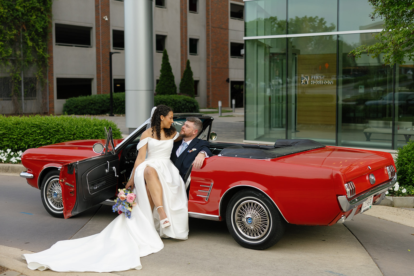 Bride sits on the edge of a red vintage Mustang as the groom wraps an arm around her during a romantic moment, taken by a Greenville SC Wedding Photographer.