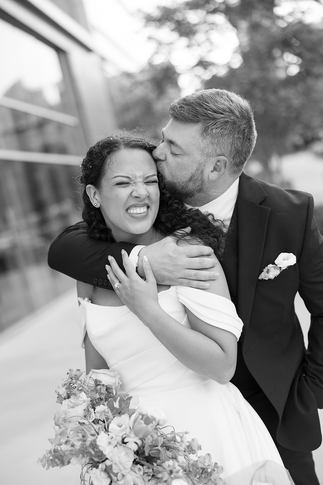 Groom kisses bride on the temple as she laughs, holding her vibrant bouquet in a joyful wedding day embrace, taken by a Greenville SC Wedding Photographer.