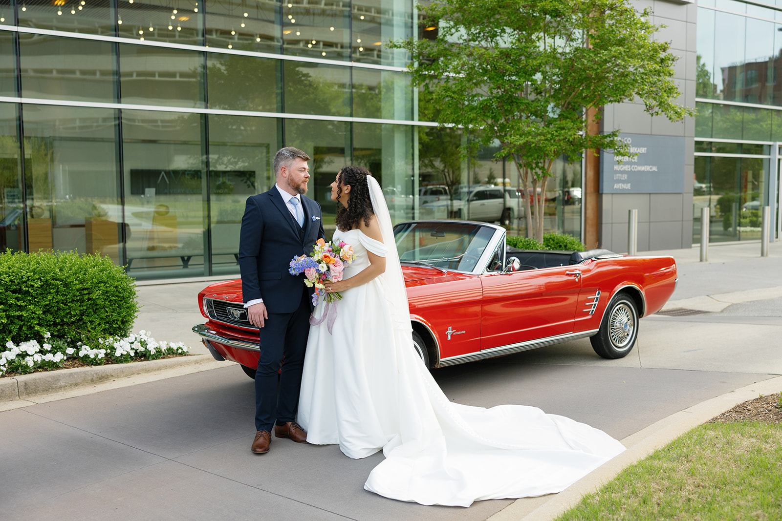 Groom embraces bride in front of a red Mustang, her elegant train spilling onto the pavement outside the venue taken by a Greenville SC Wedding Photographer.