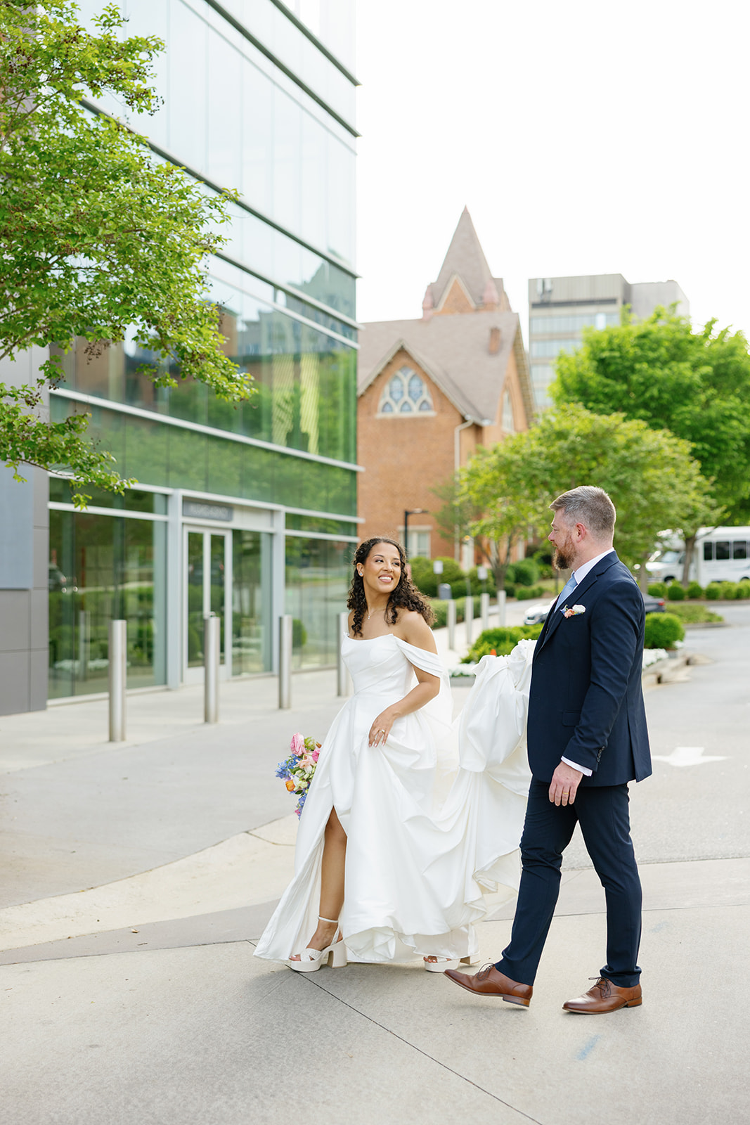 Bride and groom walk joyfully through downtown Greenville, SC, with the groom holding her timeless white train.