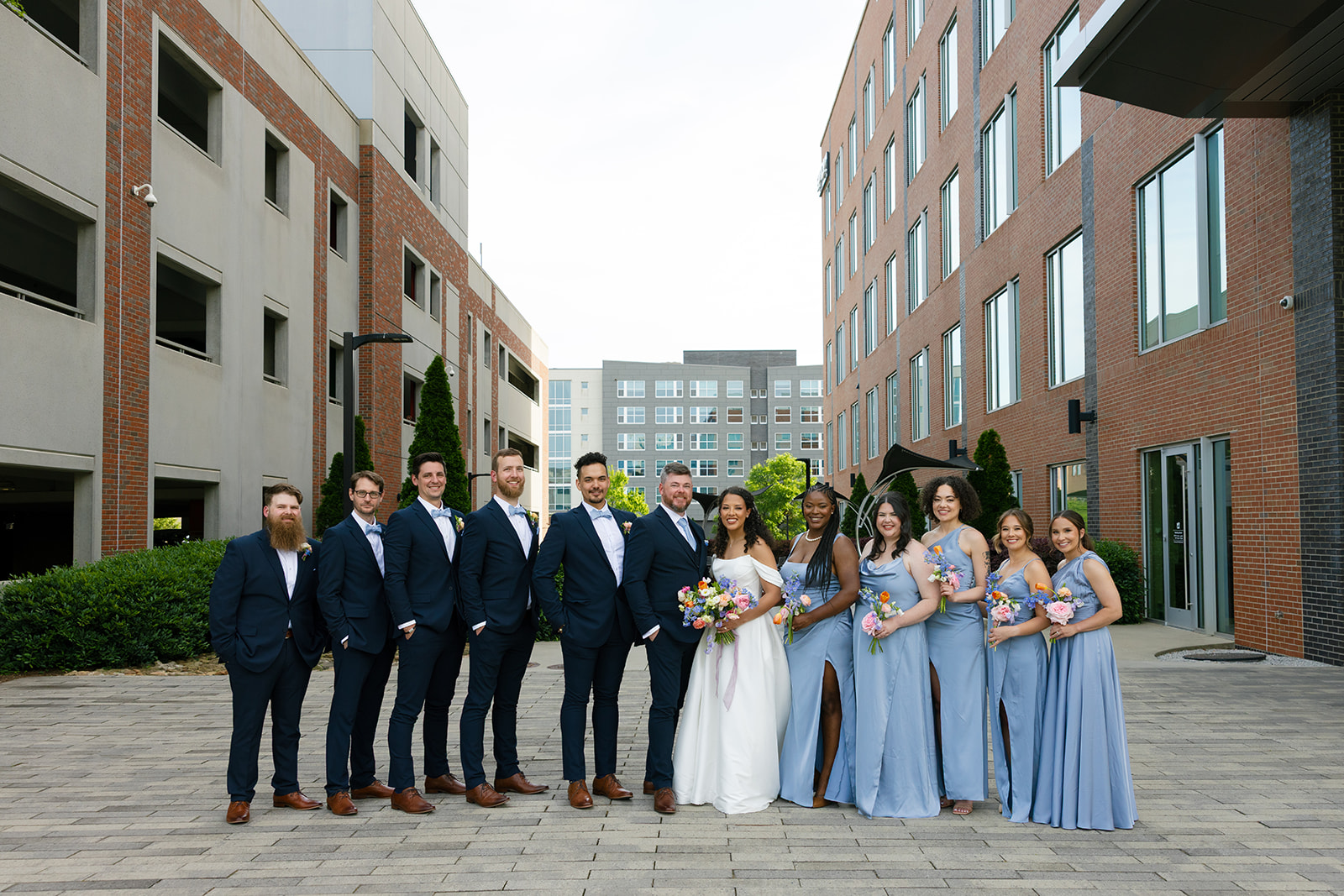 The full wedding party poses together in an urban courtyard, dressed in soft blue and navy tones.