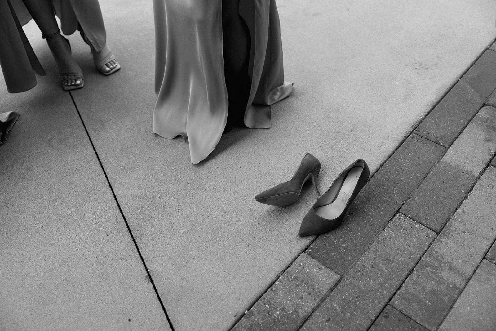 A pair of heels rest on the pavement beside bridesmaids in flowing dresses during a break from wedding festivities.
