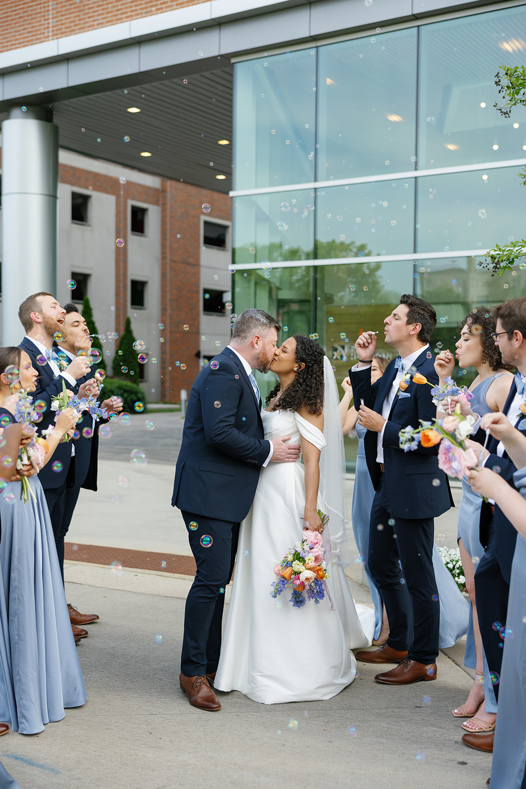 Bride and groom kiss as wedding party surrounds them blowing bubbles during a cheerful sendoff.