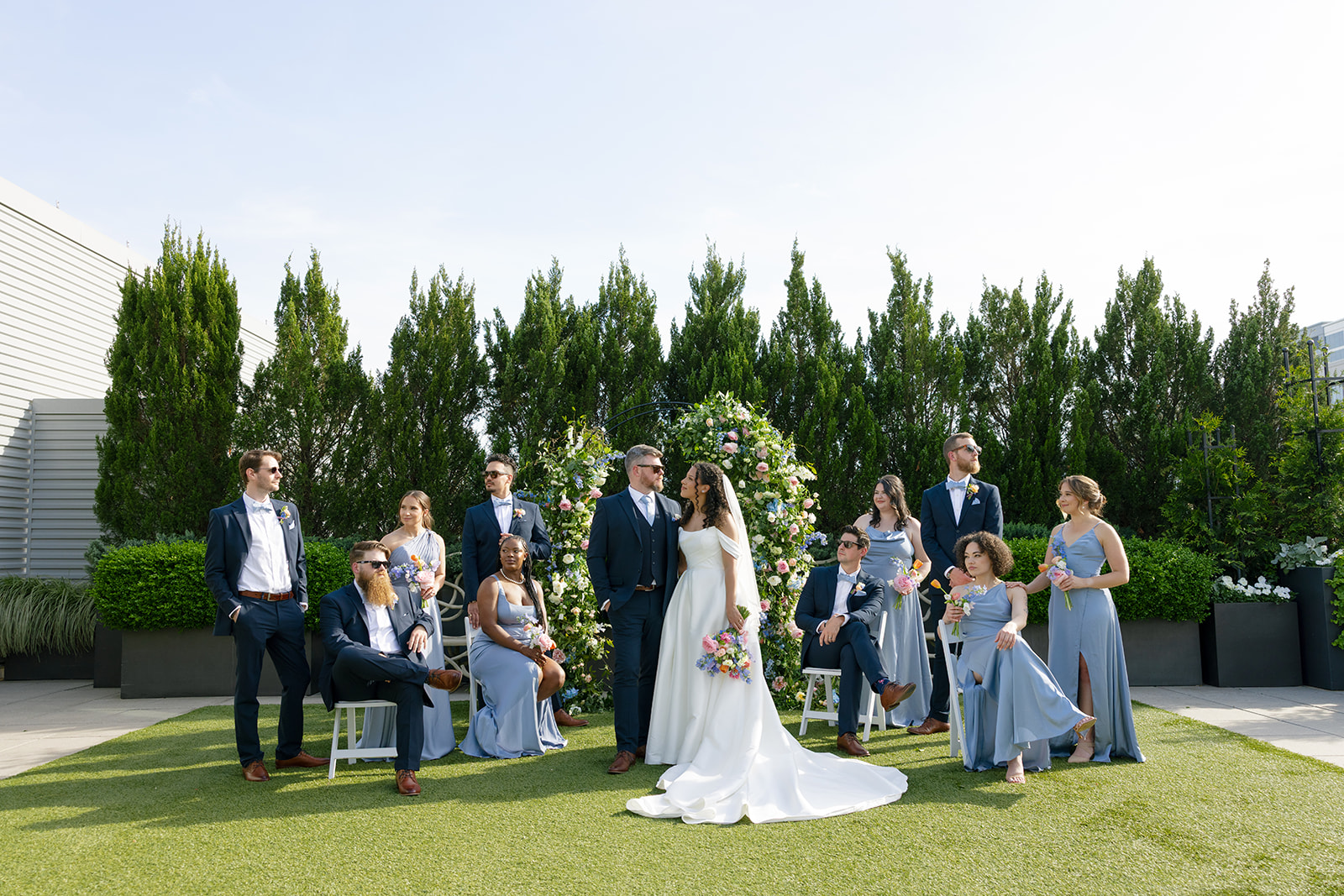 The newlyweds share a joyful moment with their wedding party in front of a lush floral ceremony arch.