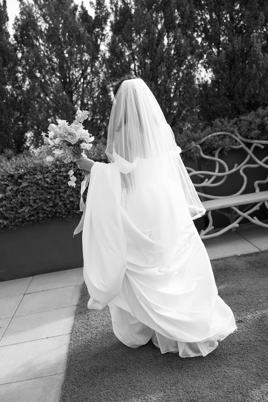 Black-and-white photo of the bride walking with her bouquet and veil trailing behind on the rooftop lawn.