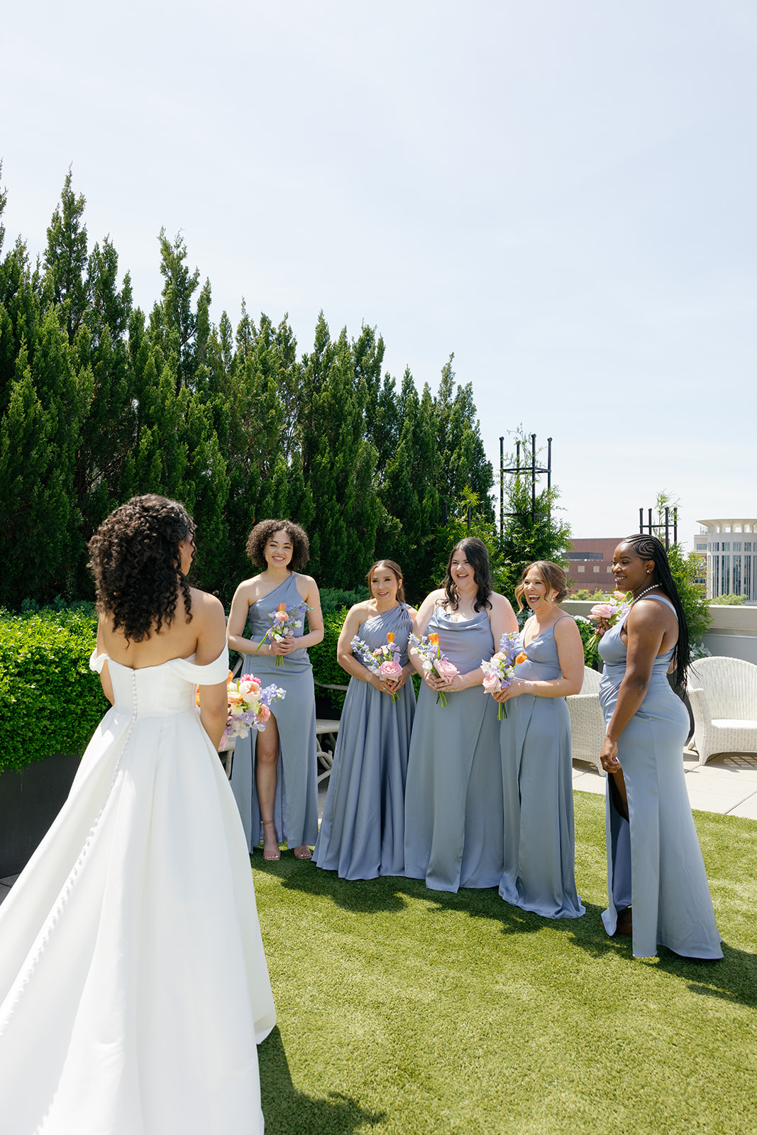 Bride approaches her bridesmaids, who react with joyful surprise during the first look on the rooftop, taken by a Greenville SC Wedding Photographer..