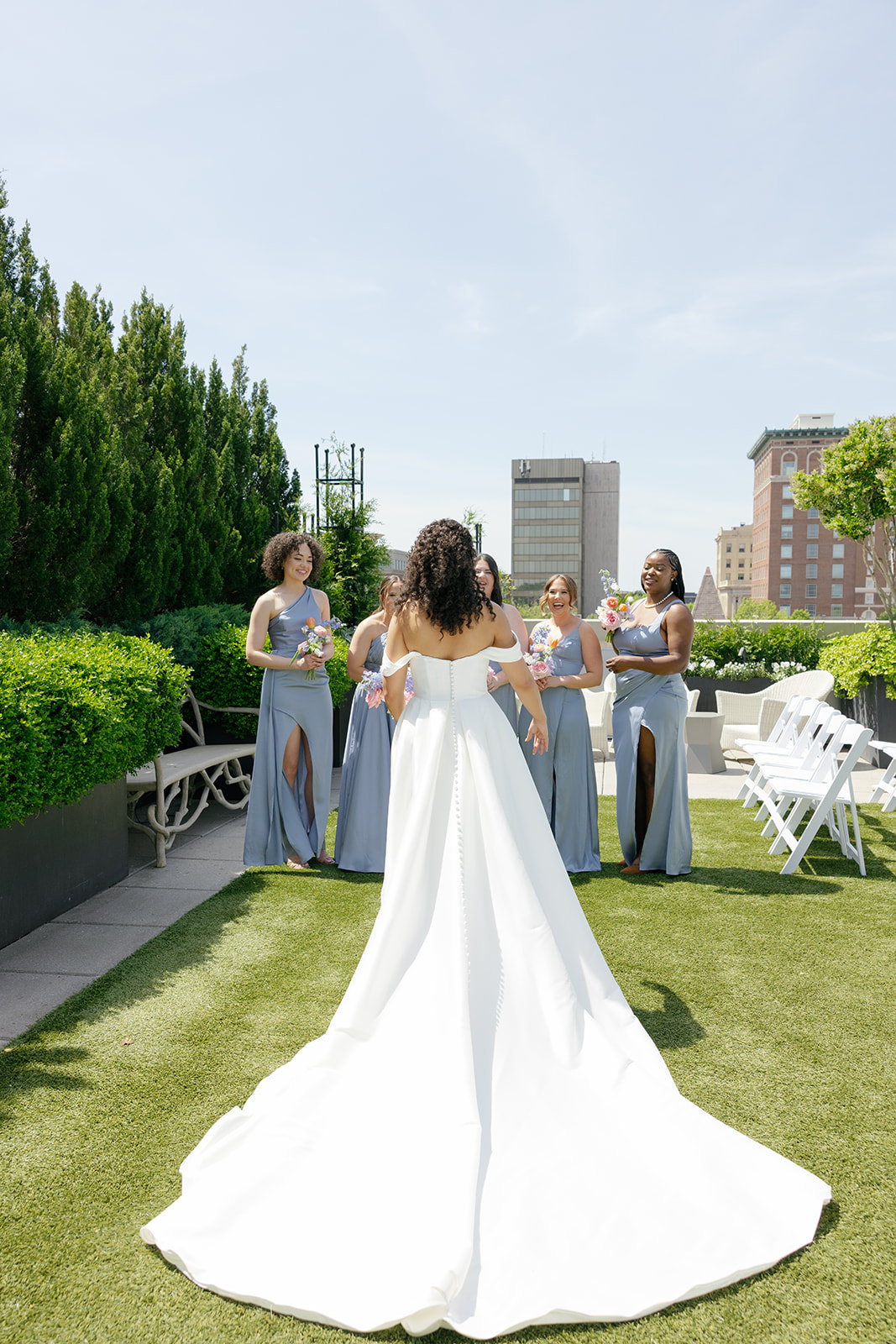 Bride walks across the rooftop lawn toward her bridesmaids in soft blue gowns, dress flowing behind her.