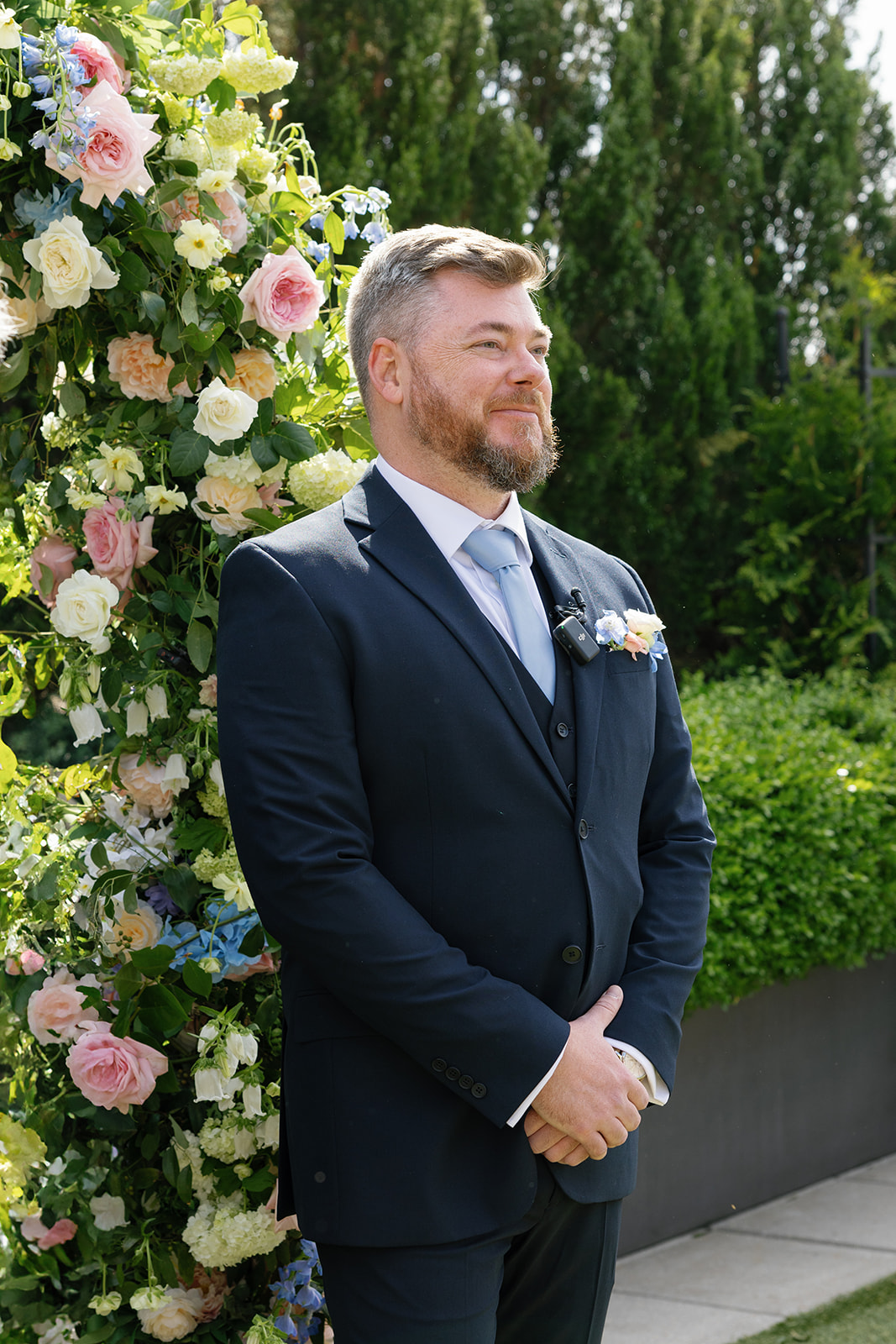 Groom smiles emotionally as he waits beneath a lush floral arch for his bride during their outdoor ceremony, taken by a Greenville SC Wedding Photographer.
