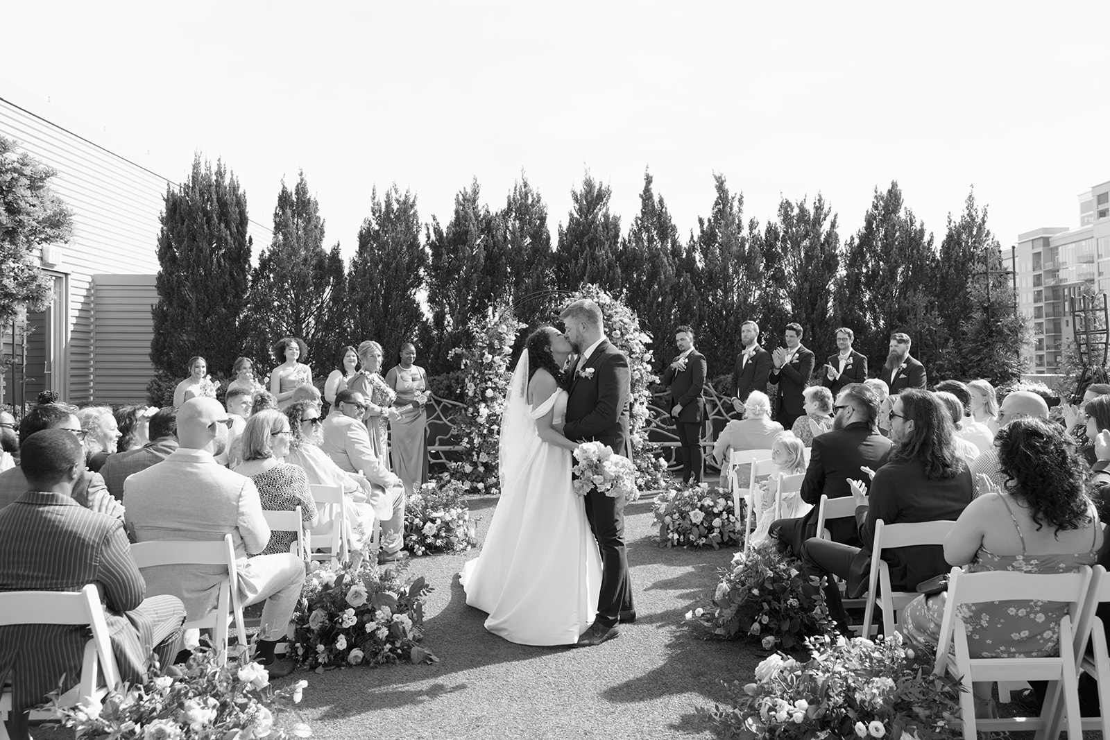 Newlyweds share a first kiss under a romantic floral arch as guests cheer during their outdoor ceremony, taken by a Greenville SC Wedding Photographer.