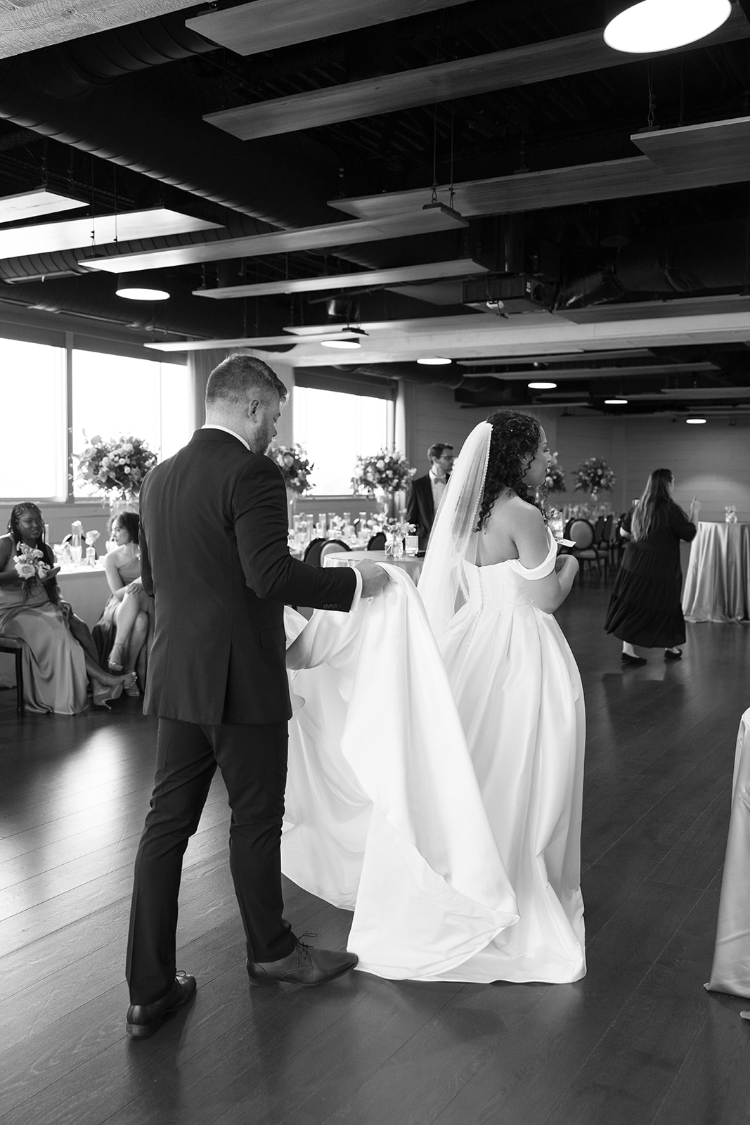 Groom gently carries the bride’s train as they walk together across the elegant indoor venue, taken by a Greenville SC Wedding Photographer.