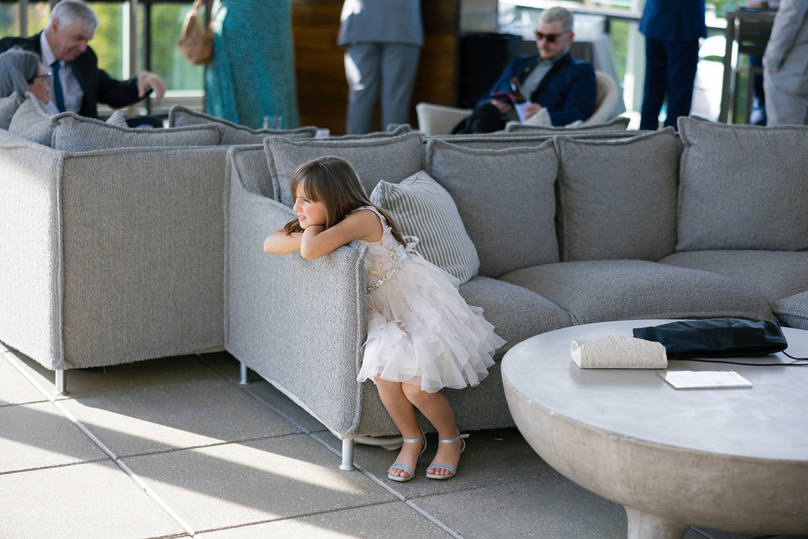 A young flower girl in a layered dress leans on a couch, watching the wedding reception unfold.