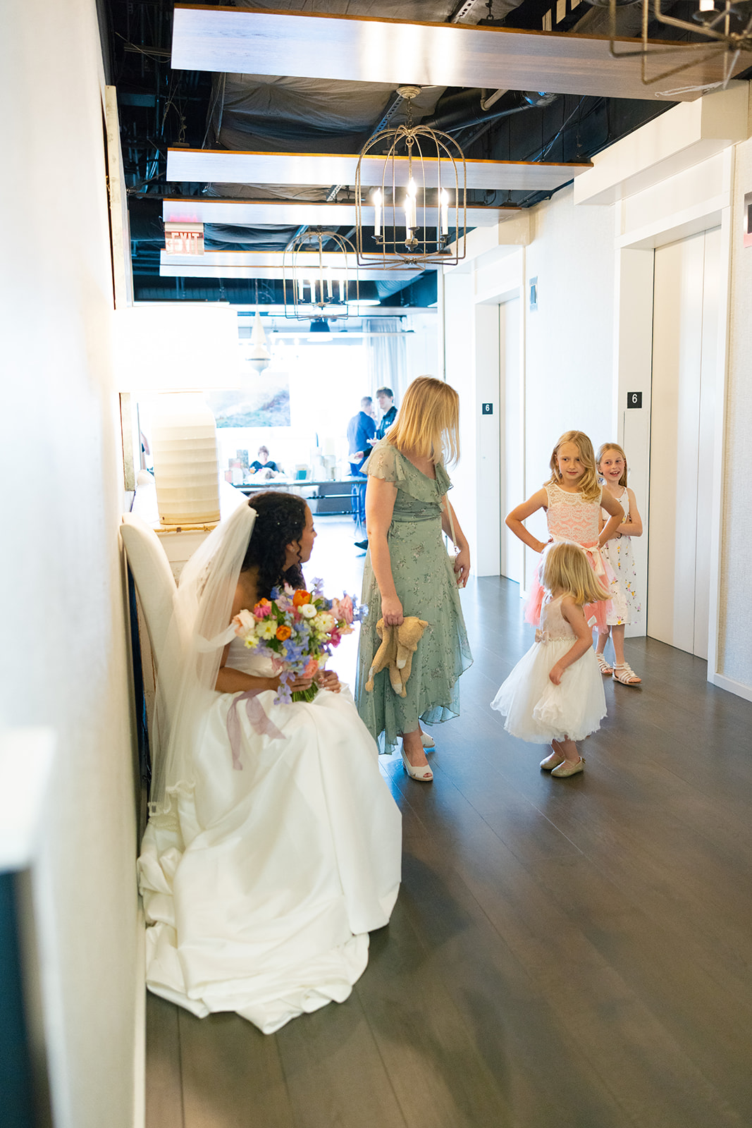 The bride sits with her bouquet, smiling at flower girls gathered around her in a quiet hallway moment.