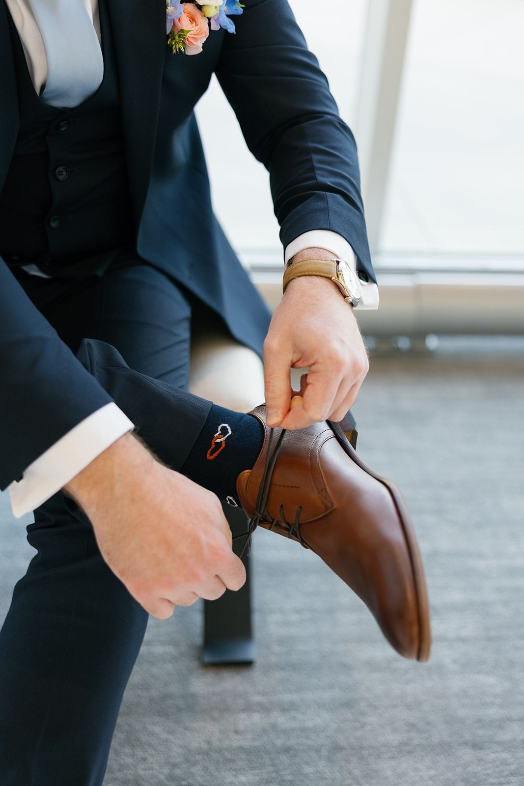 Groom ties brown leather shoes, dressed in a navy suit with playful socks and a pastel boutonnière.