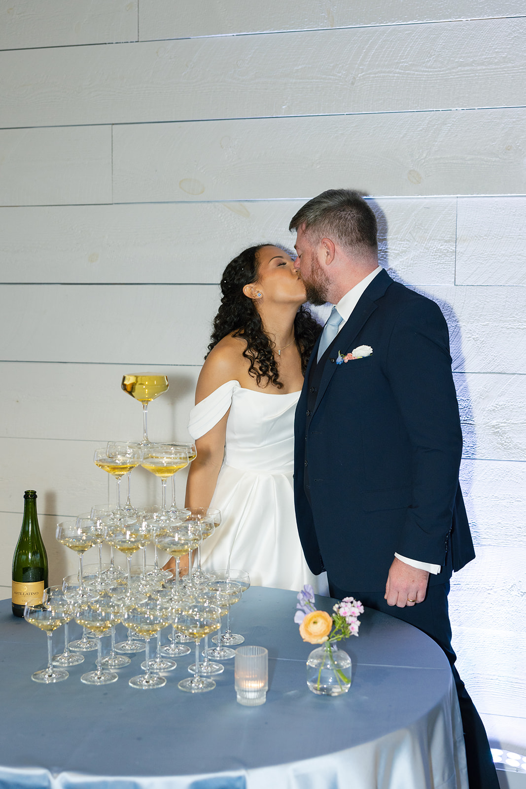 Newlyweds share a kiss while standing beside a tower of filled champagne coupes.