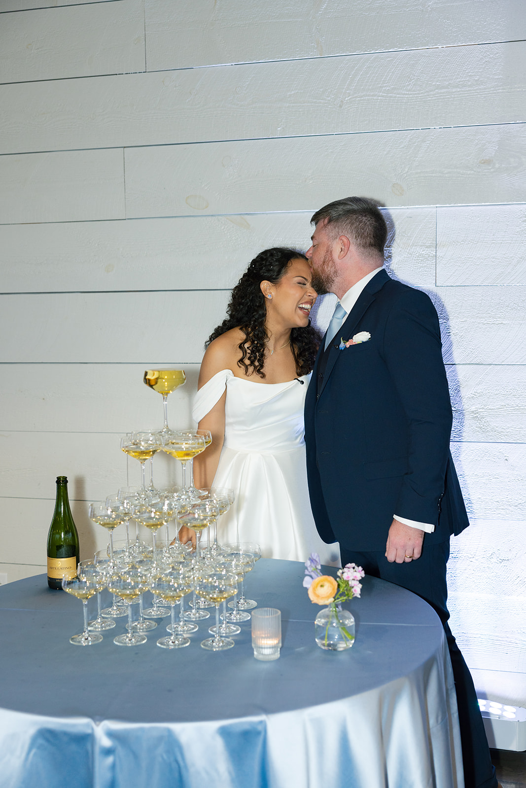 Groom kisses the bride on the forehead as they laugh beside a champagne tower, taken by a Greenville SC Wedding Photographer.
