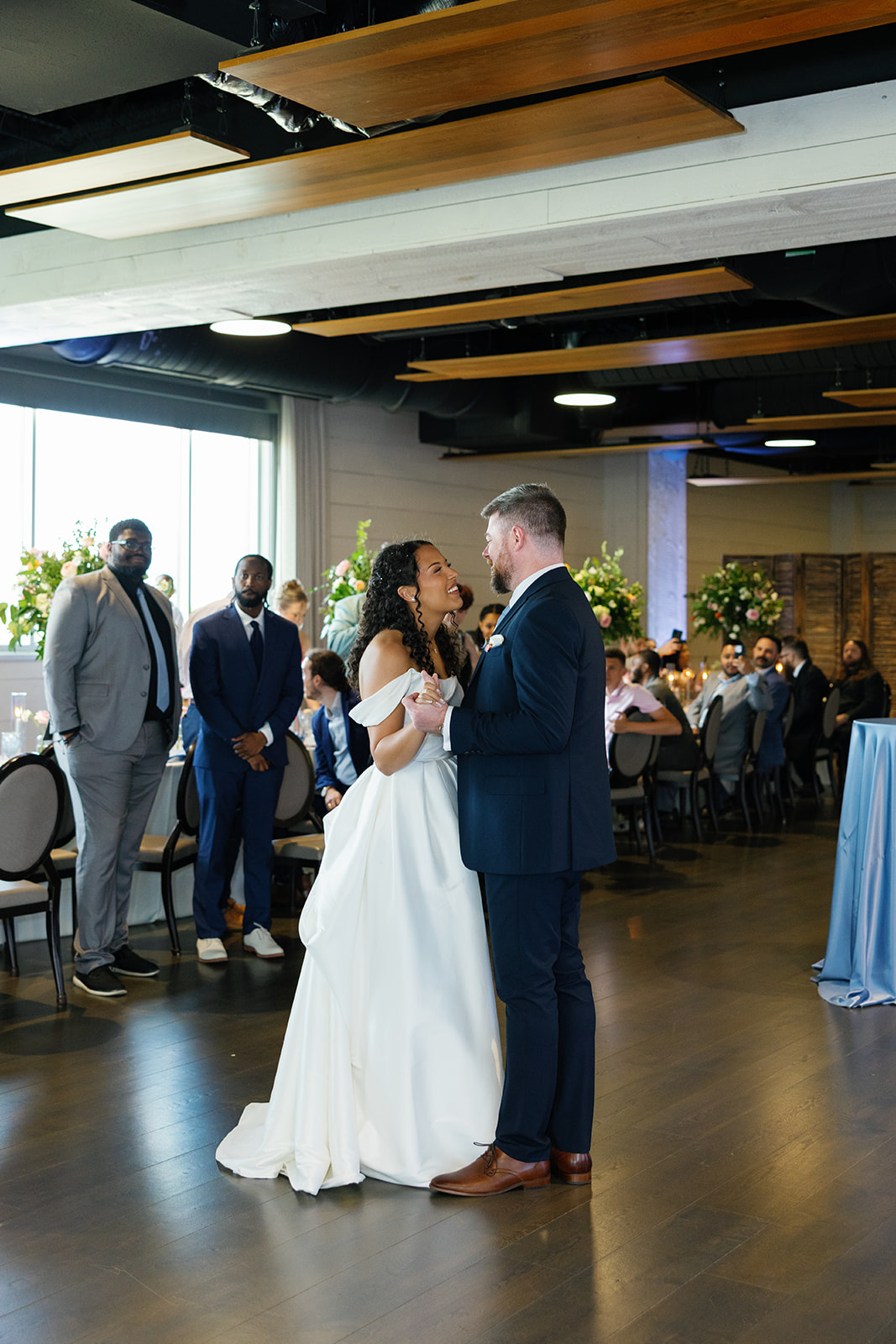 Bride and groom share a romantic first dance surrounded by guests and soft floral decor.