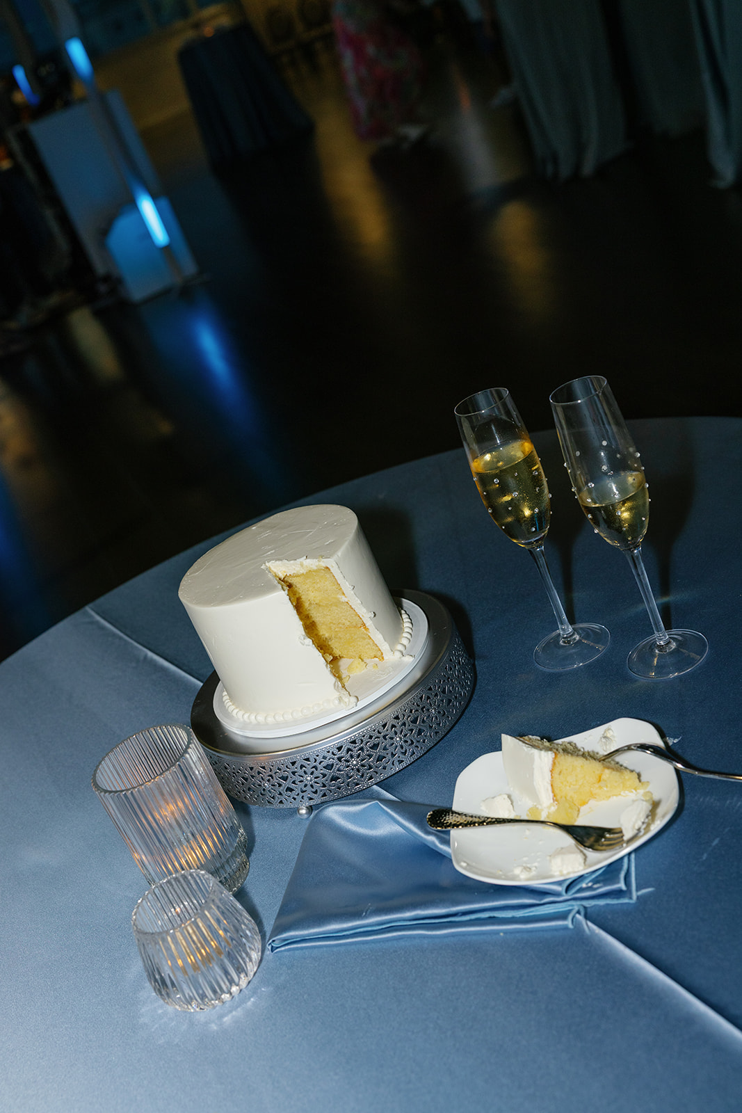 A minimalist white wedding cake with two champagne flutes sits on a blue satin table after the cake cutting.