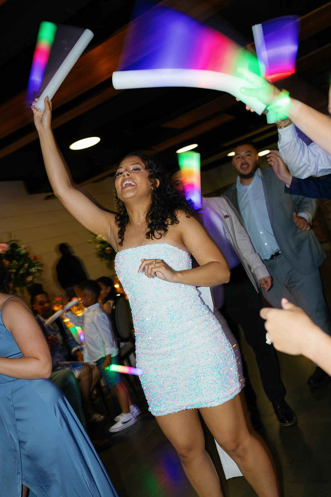 Bride dances under colorful lights holding glow sticks during a vibrant wedding party.