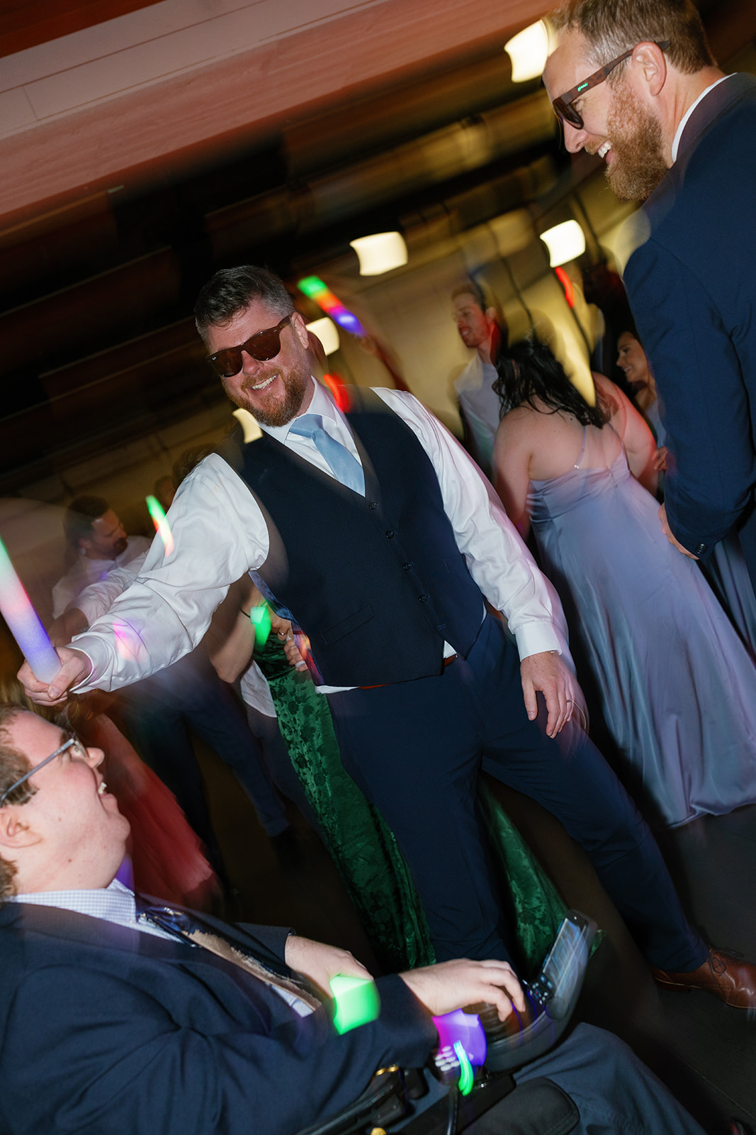 Groom dances with friends, grinning behind sunglasses and waving a glowing light stick at the reception.