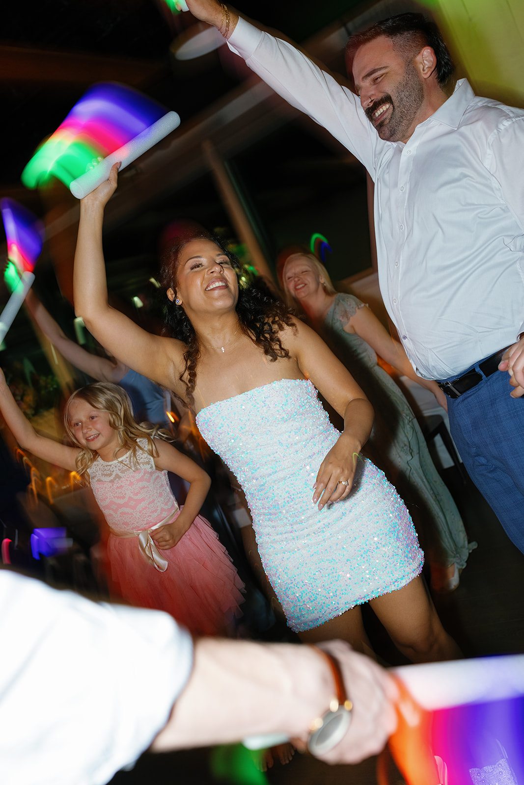 Bride and guests dance under glowing lights, waving neon foam sticks in a lively, joyful celebration.