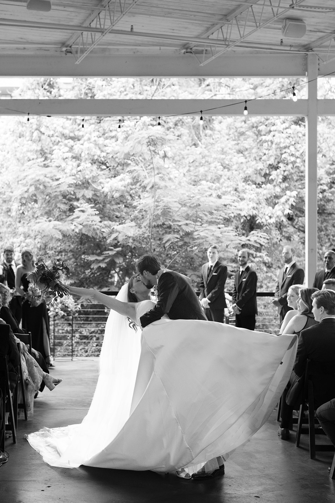 Groom dips and kisses the bride at the end of their ceremony as guests cheer beneath a canopy of greenery.