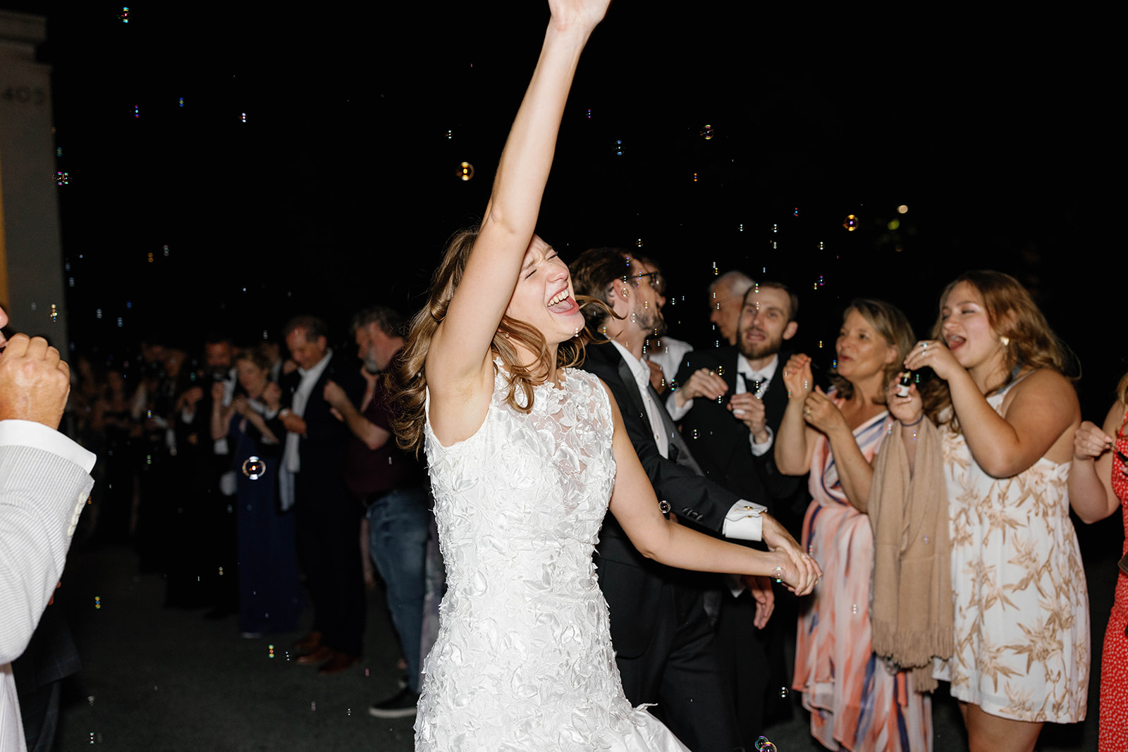Bride throws her arm up in celebration during a bubble exit with guests gathered around the venue steps.