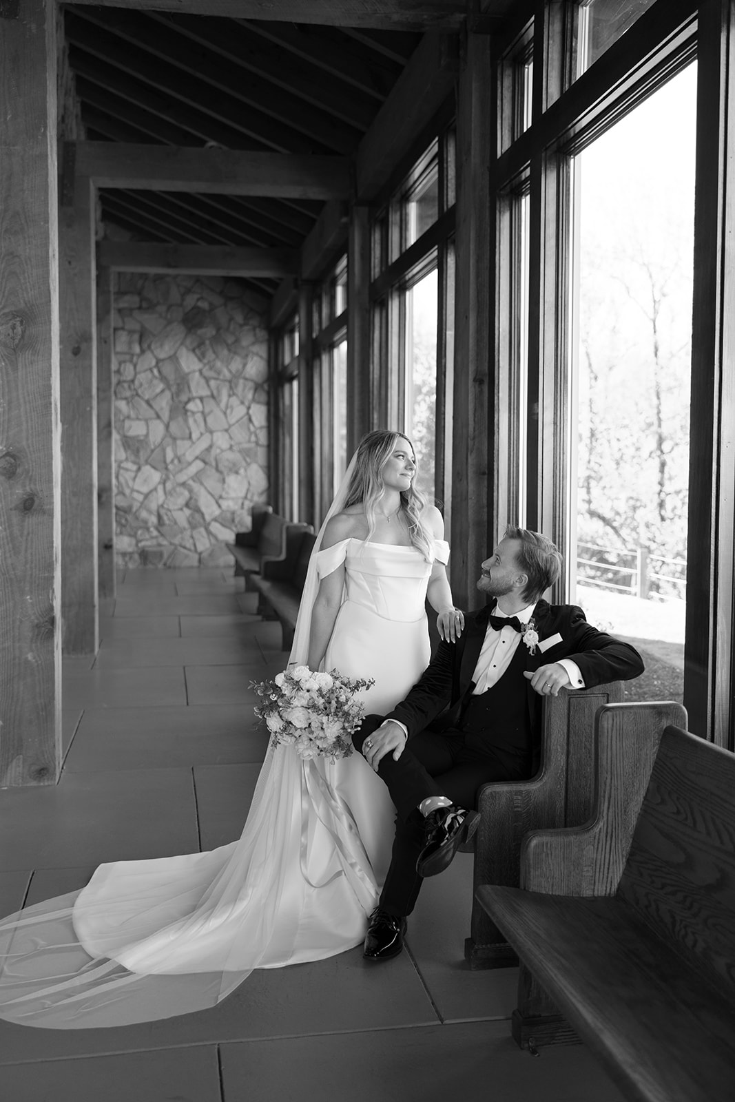 bride looking through window of Glassy Chapel while groom smiles up at her