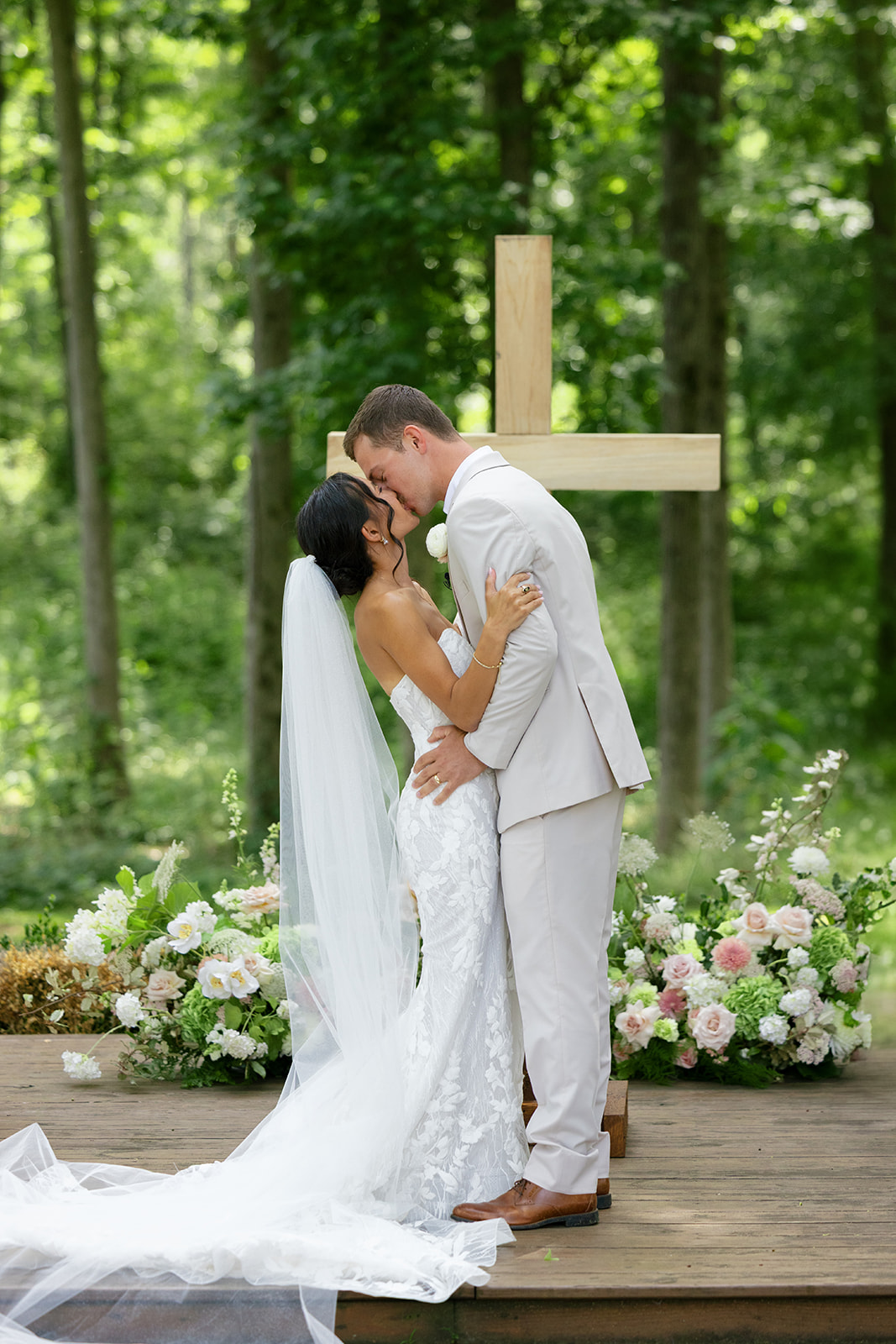 bride and groom share a kiss after exchanging their wedding vows in front of a large wooden cross surrounded by florals