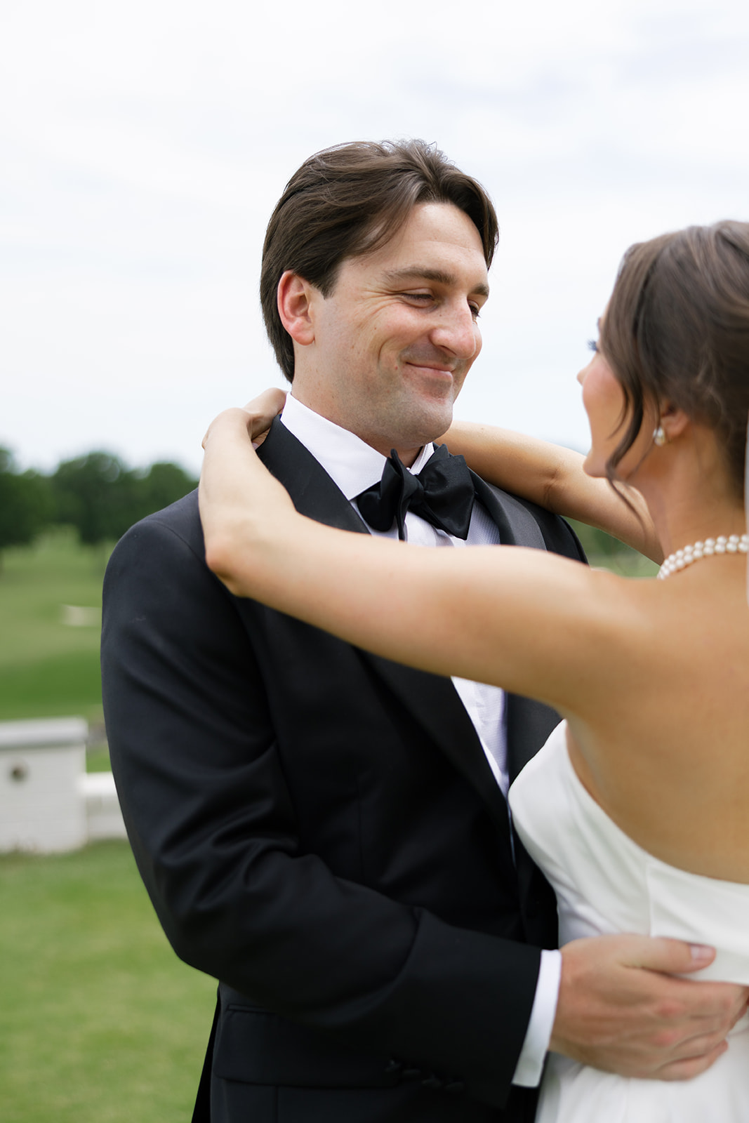 Groom smiling with pride while bride wraps her arms around his shoulders, outdoors on the golf course.