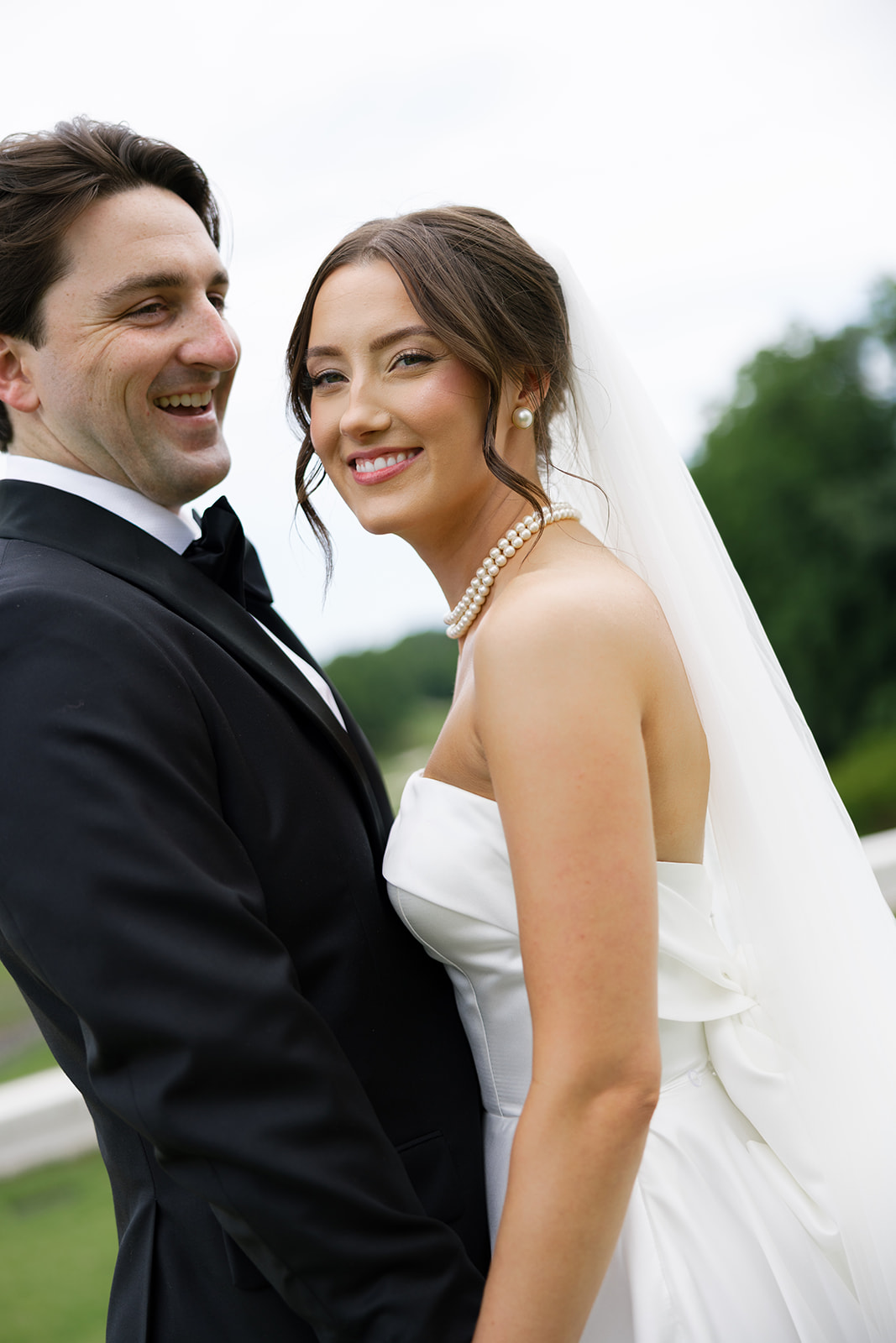 Bride and groom smiling closely together, bride in strapless gown with pearls and veil, groom in black tuxedo.