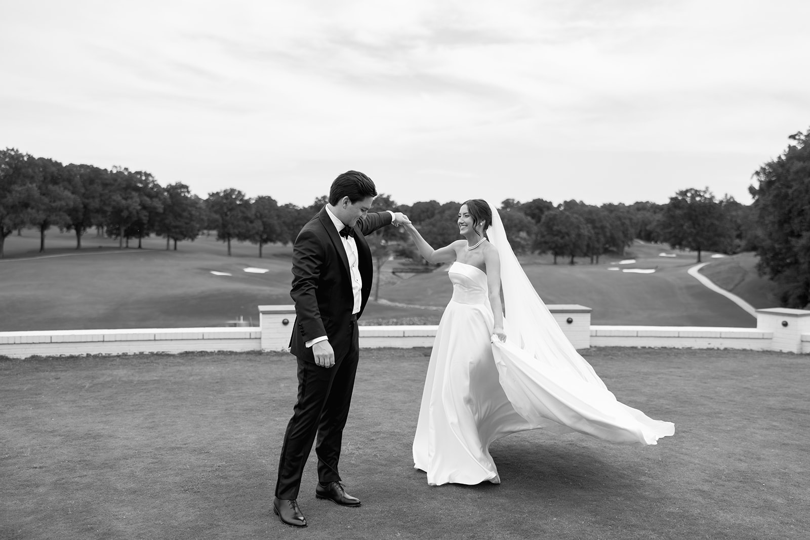 Black-and-white photo of the groom spinning the bride playfully on the golf course, her dress and veil sweeping the ground.
