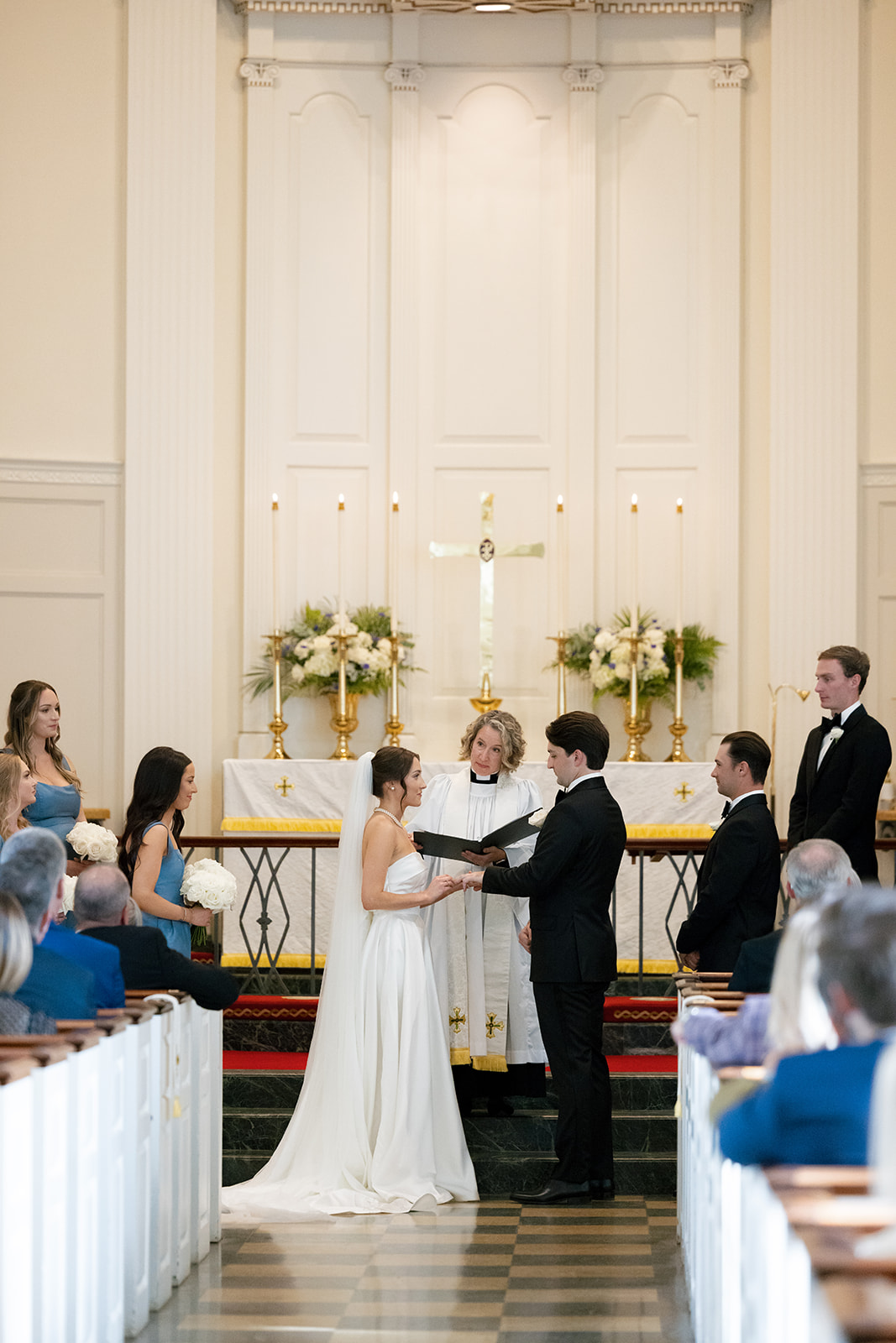 he bride and groom face each other holding hands as the officiant leads them through vows at Christ Episcopal Church, surrounded by bridesmaids and groomsmen.