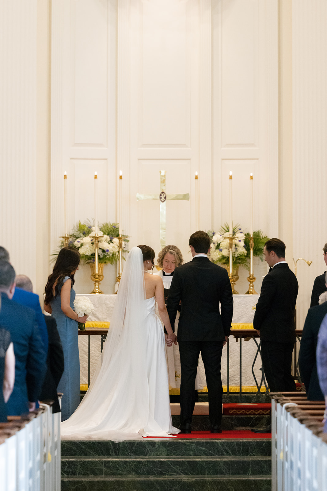 The couple stands hand in hand with heads bowed as the officiant leads a prayer in front of the golden cross and altar adorned with white flowers.
