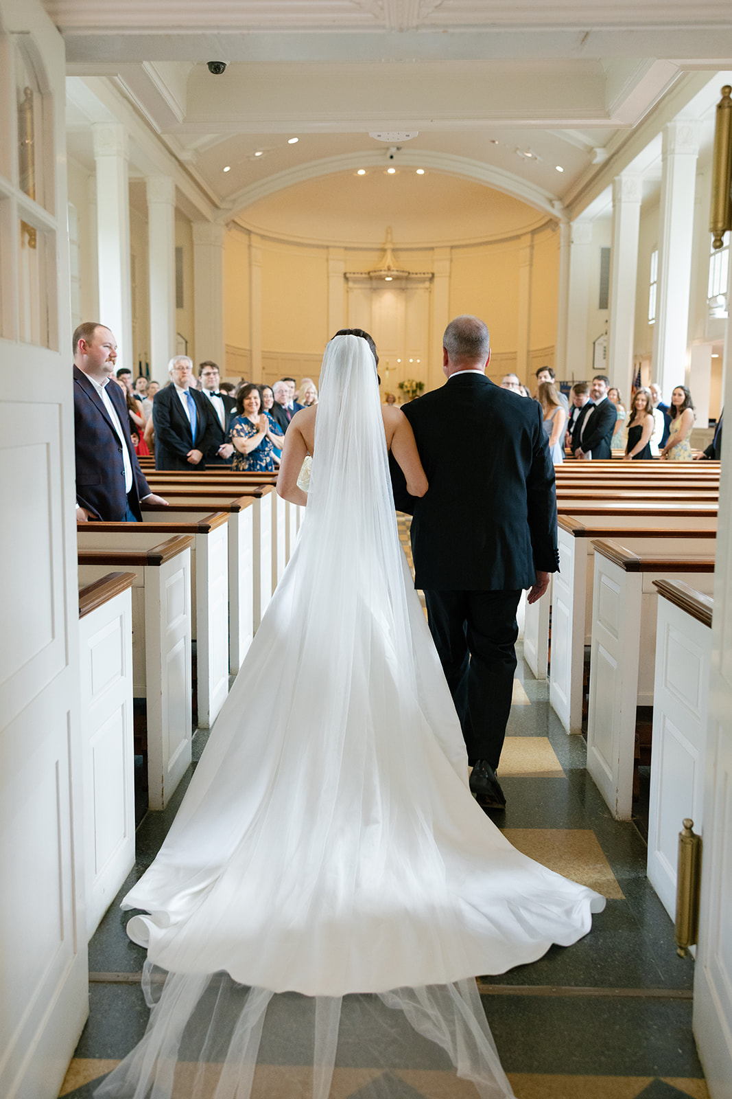 A bride in a flowing white gown and long veil walks arm in arm with her father down the aisle of Christ Episcopal Church, with wedding guests smiling and watching.