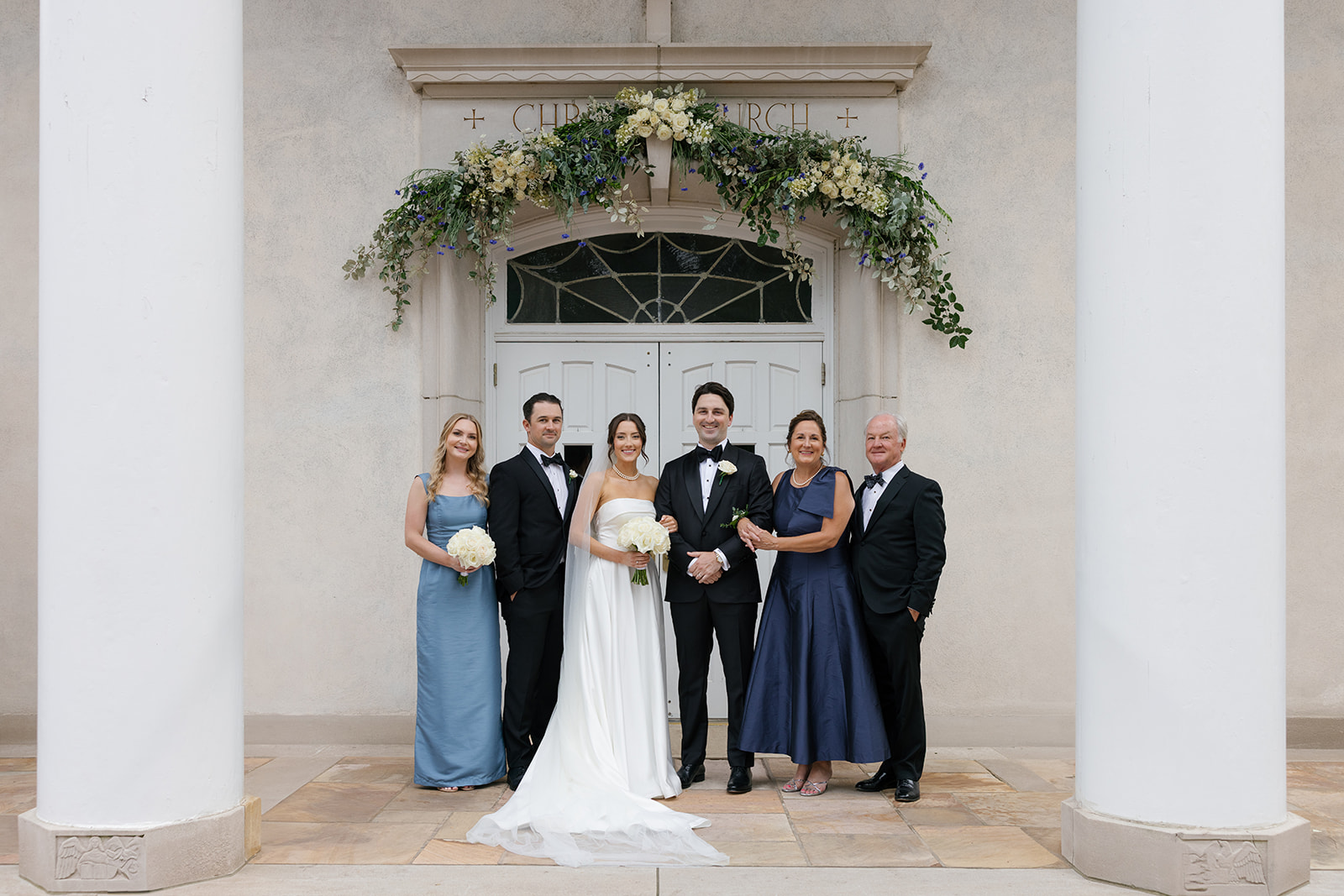 Formal family portrait in front of the church doors with the bride and groom surrounded by parents and siblings, beneath a floral garland arch.