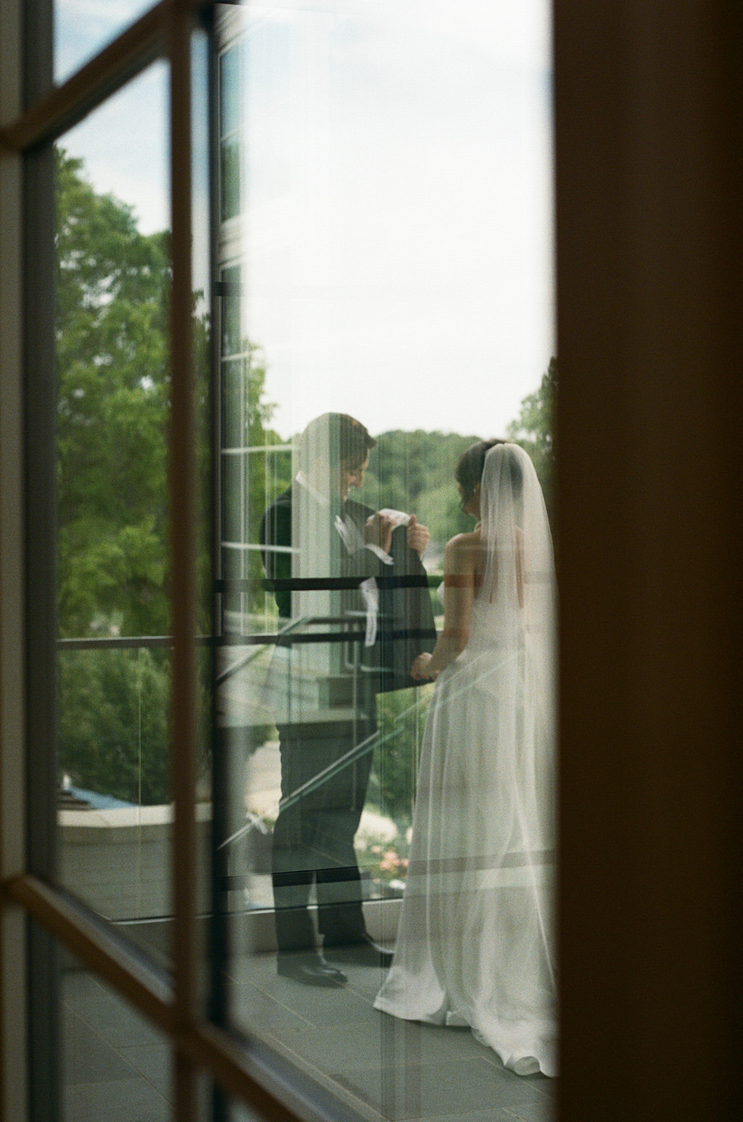 Dreamy film photo of bride and groom sharing a private moment on a balcony, captured through a window reflection.