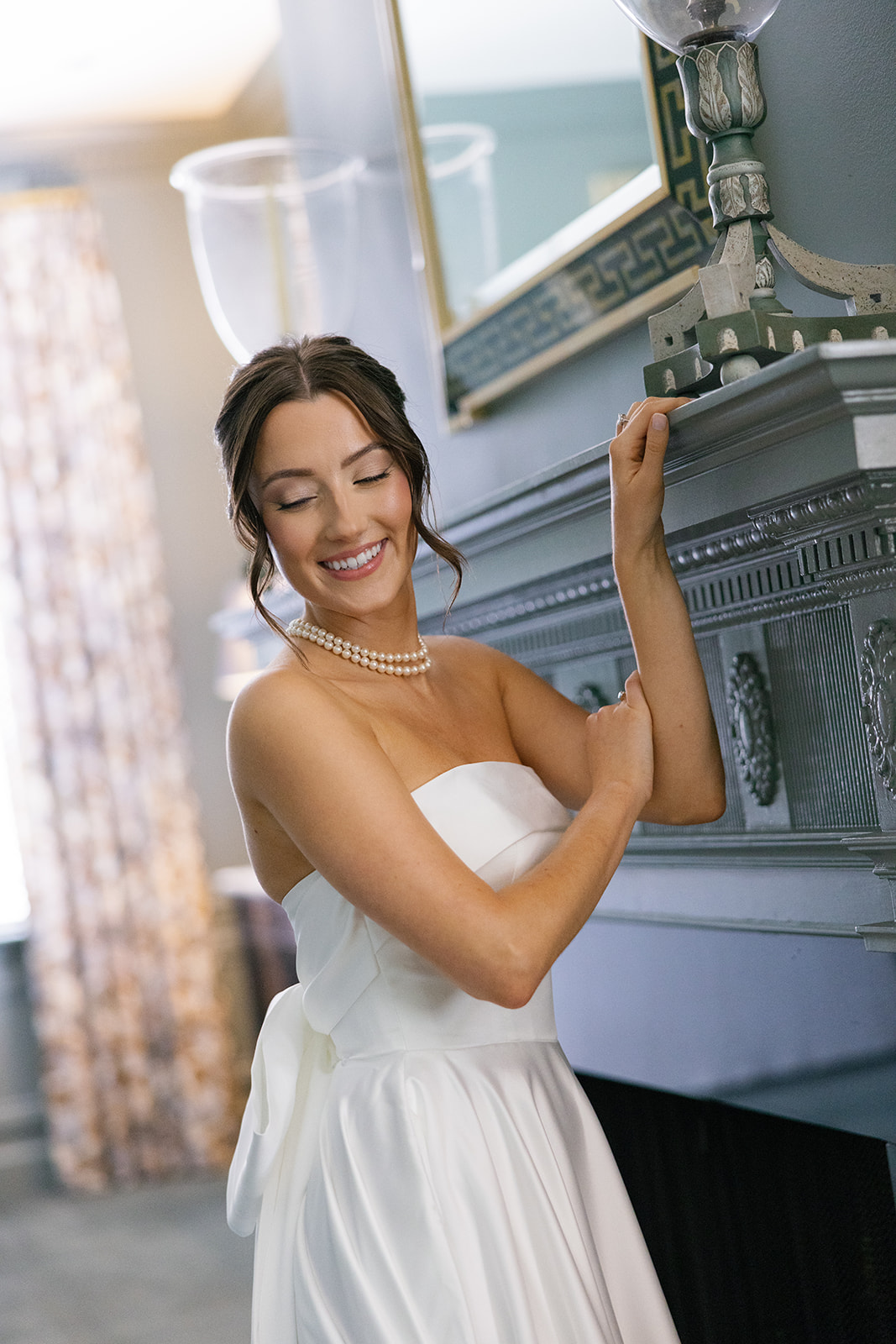 Bride in strapless white gown with pearl necklace smiling softly while resting her arm on a fireplace mantle.