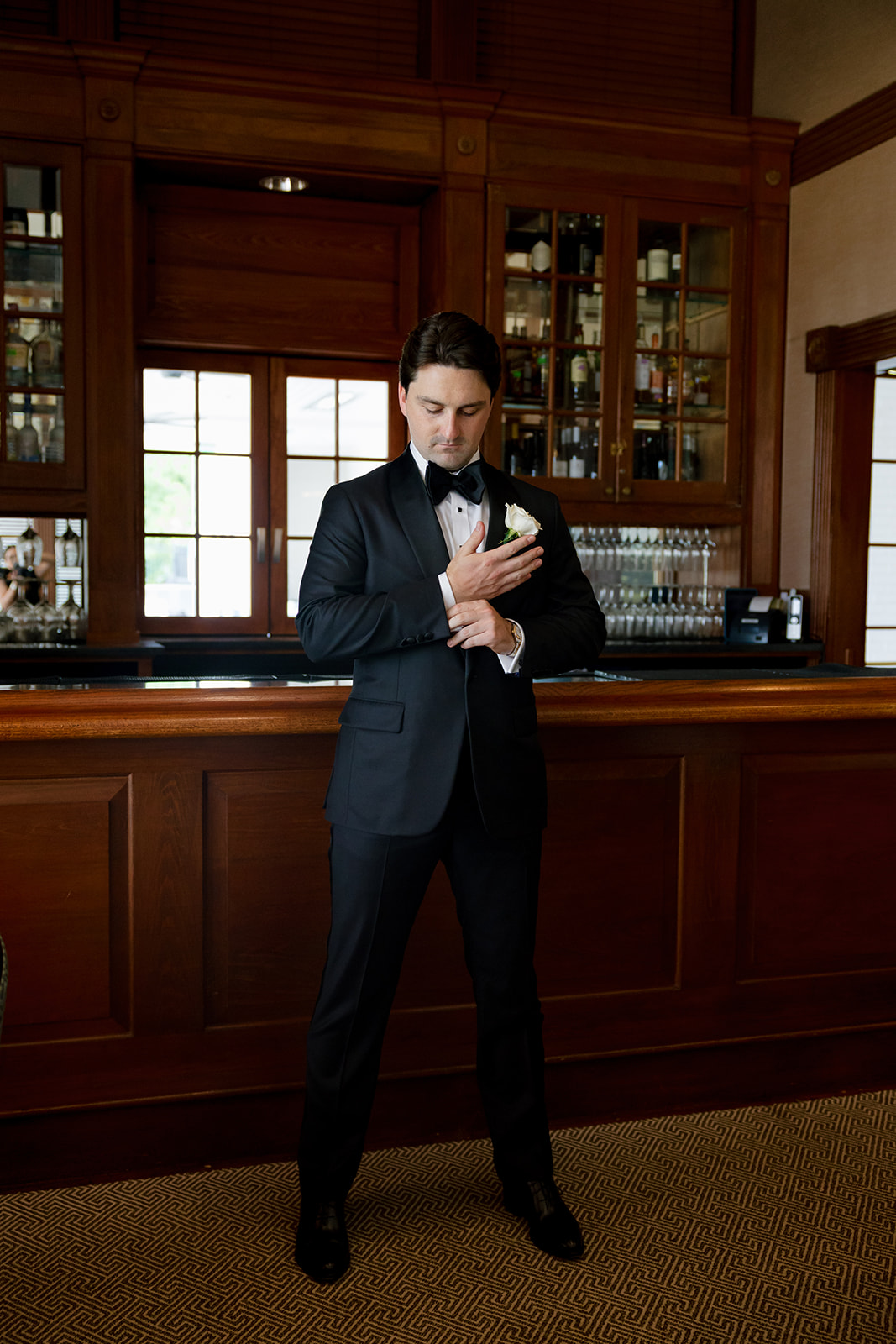 Groom adjusting his cufflinks in a black tuxedo with a white rose boutonniere at Myers Park Country Club.