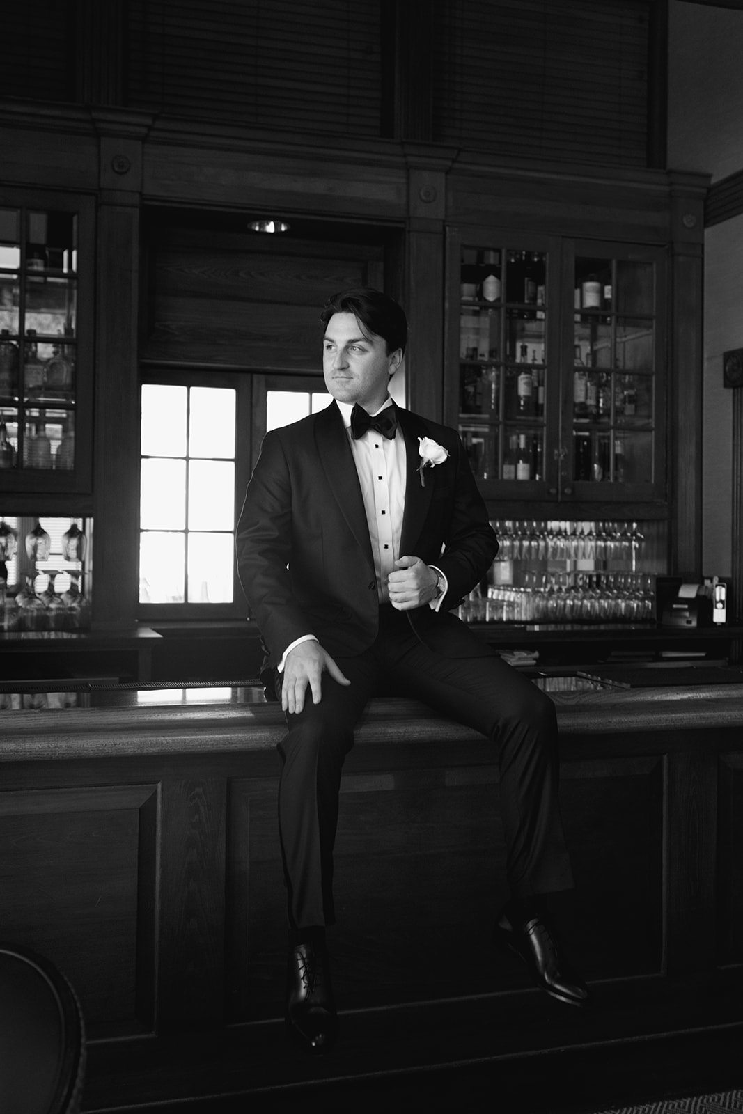 Black and white portrait of the groom sitting casually on the bar, looking off to the side in his tuxedo.