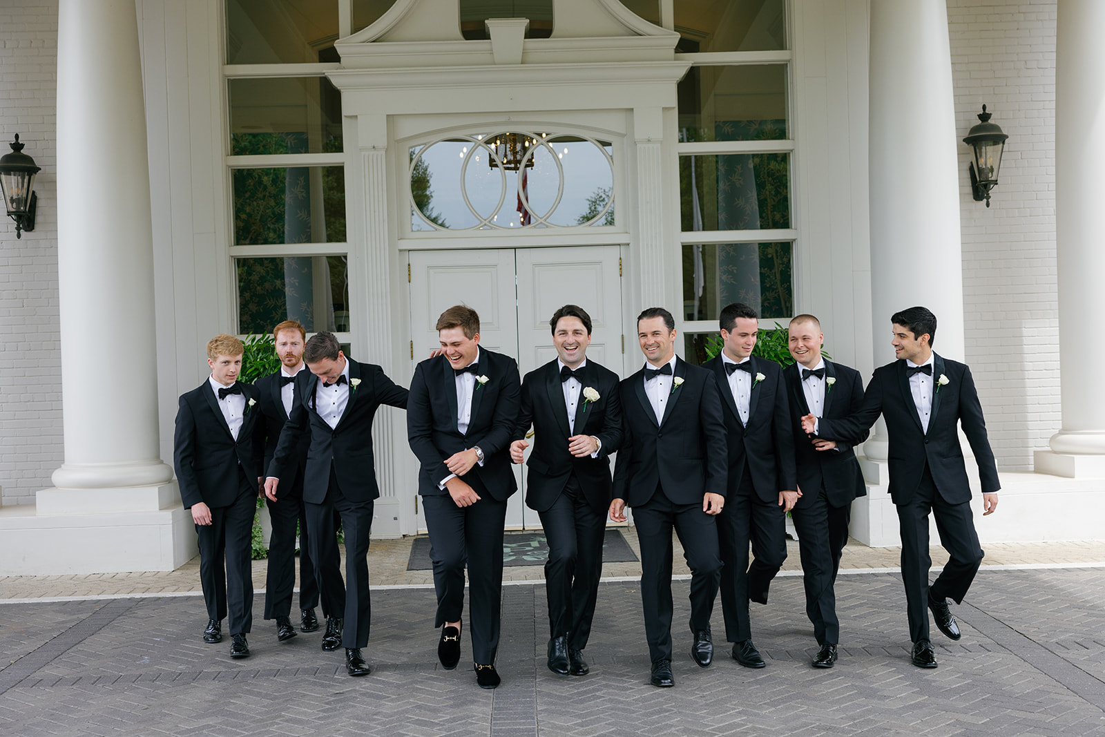 Groom and groomsmen in black tuxedos walking together outside the venue, laughing.