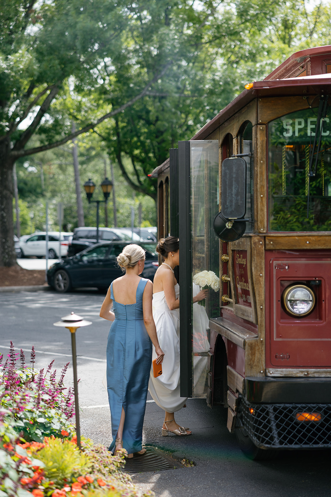 Bride stepping onto a vintage-style trolley bus, bouquet in hand, with a bridesmaid in a blue gown following behind her.