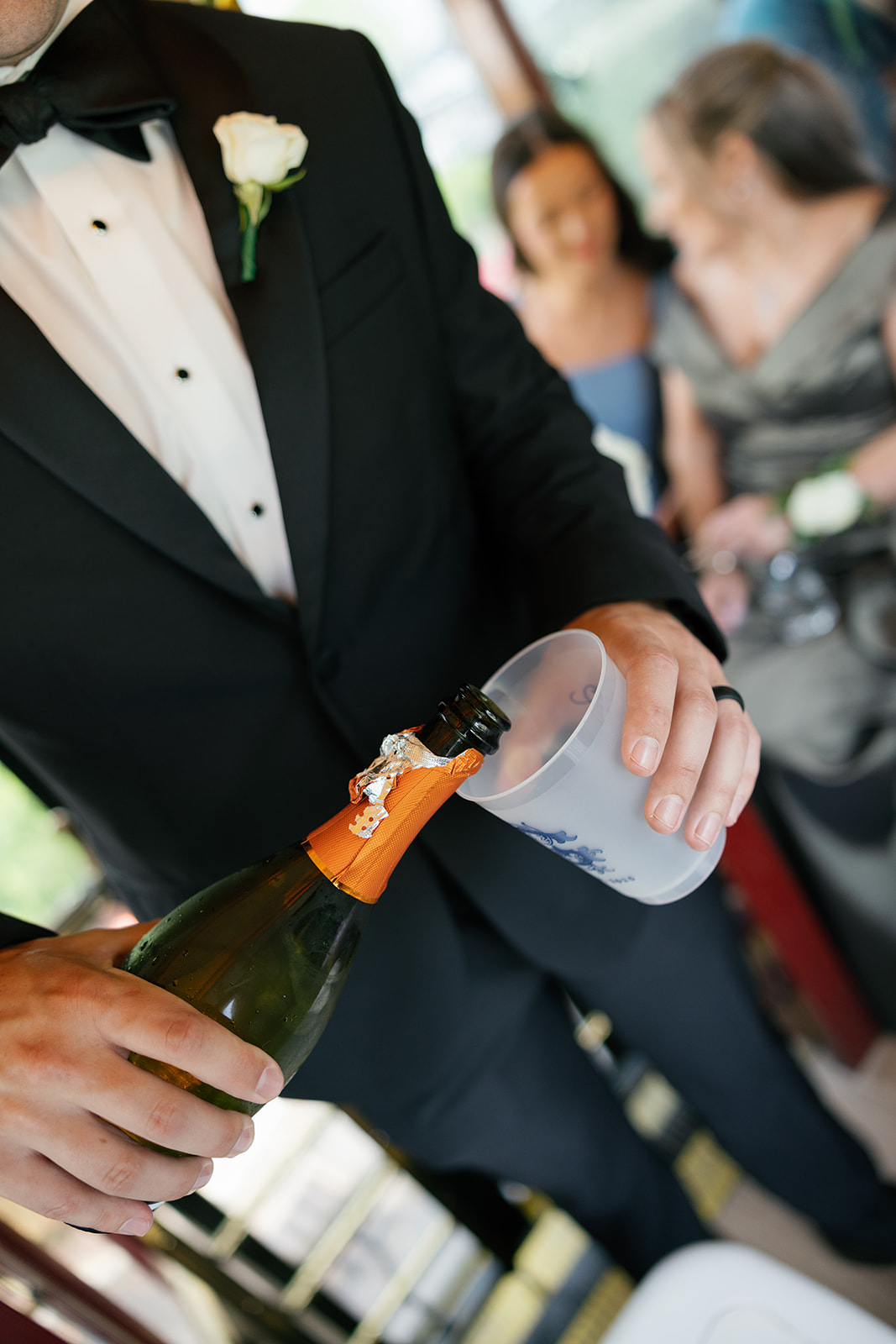 Groom in a black tuxedo pouring champagne into a plastic cup while on a trolley, with wedding guests blurred in the background.