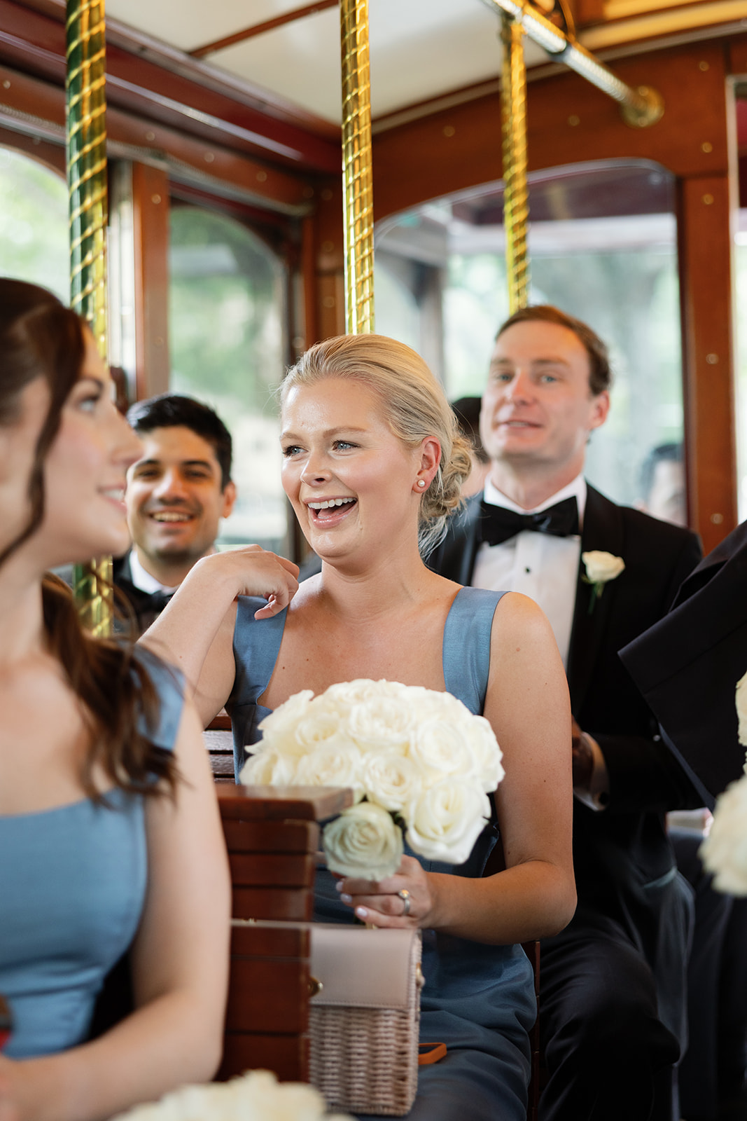 Bridesmaid in a dusty blue dress holding a white rose bouquet, laughing and chatting with the wedding party inside a trolley.