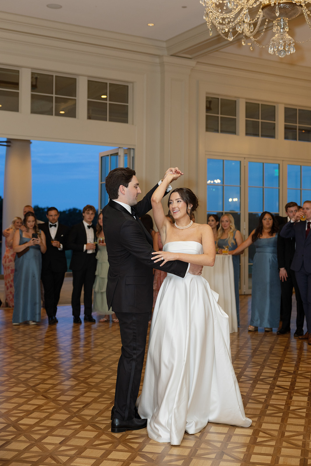 Groom twirls the bride on the dance floor during their first dance, her gown flowing as guests watch.