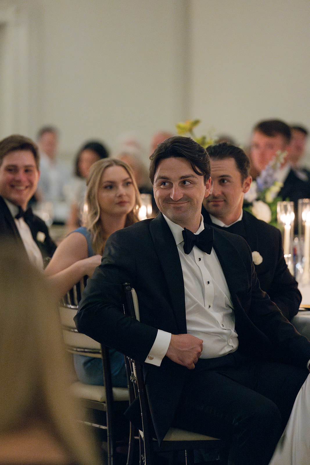 Groom grins while seated at the reception, surrounded by friends and family.