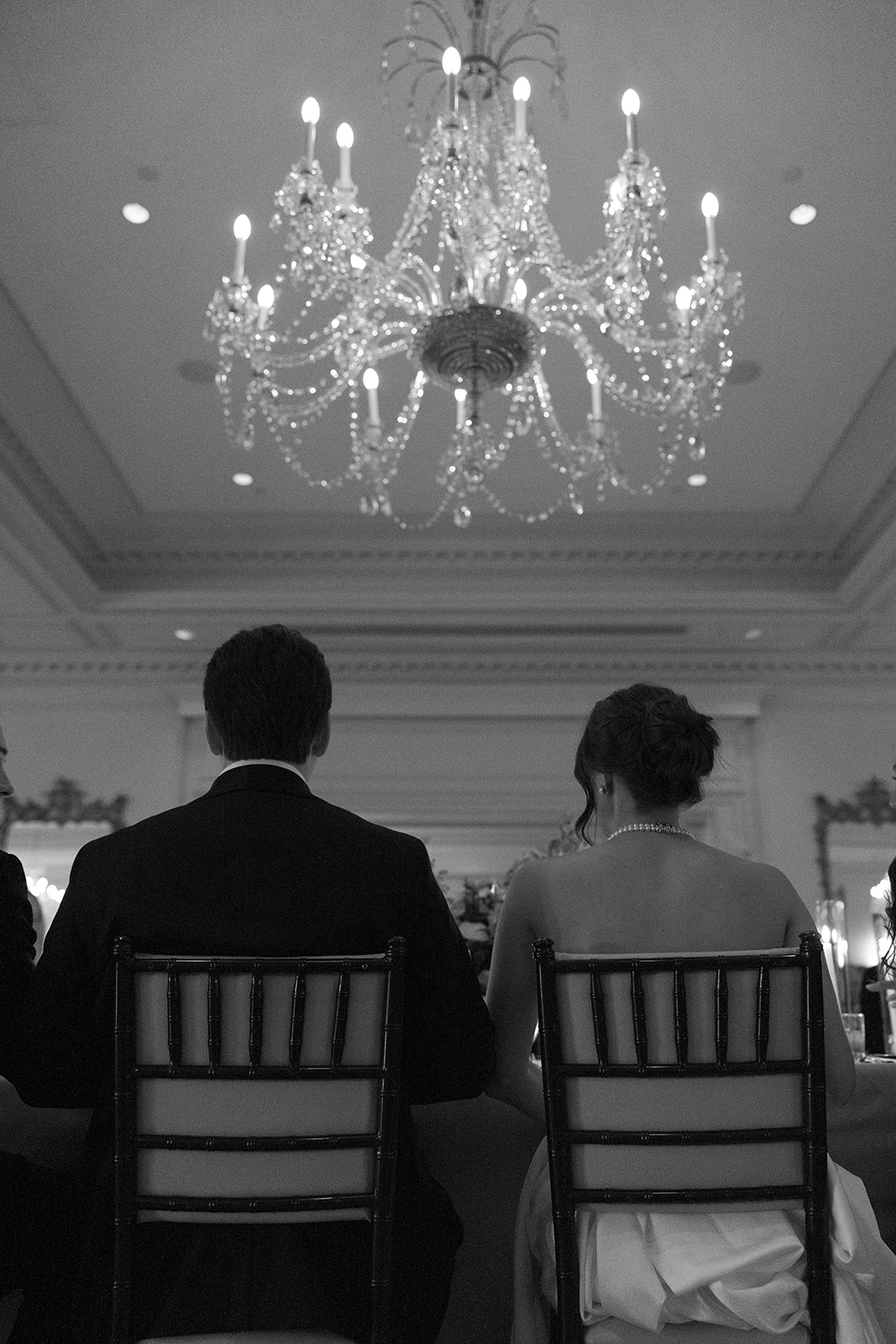 Black and white photo of the couple seated together under a grand crystal chandelier at their reception.