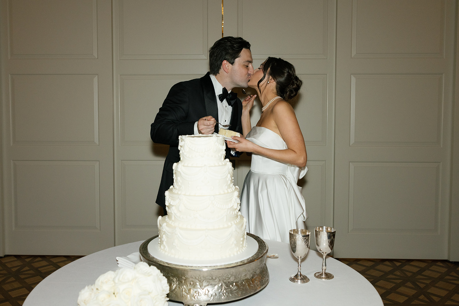 Couple kisses while holding plates of wedding cake, the cake displayed in the foreground.