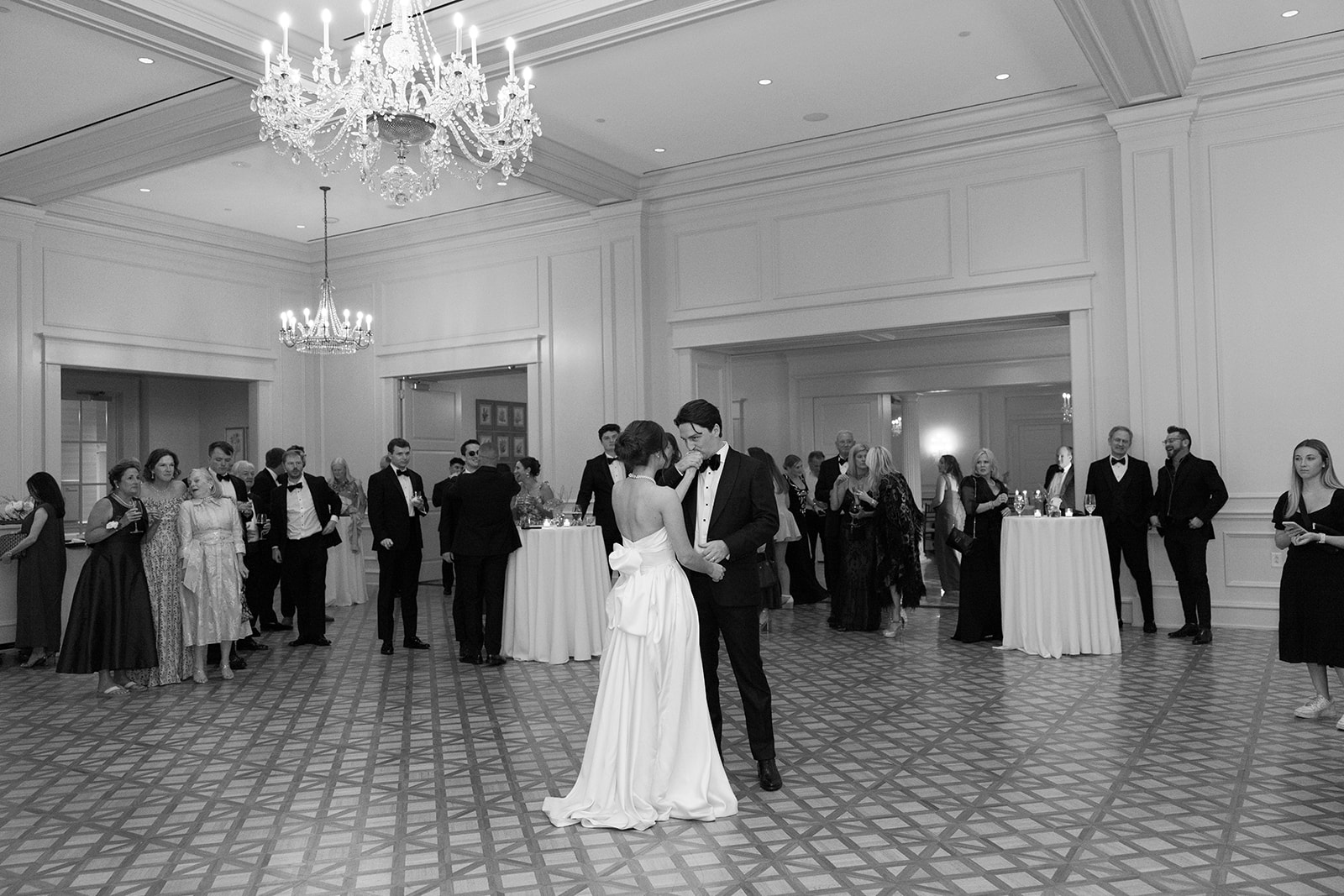 Black and white photo of the bride and groom sharing their first dance while guests look on.