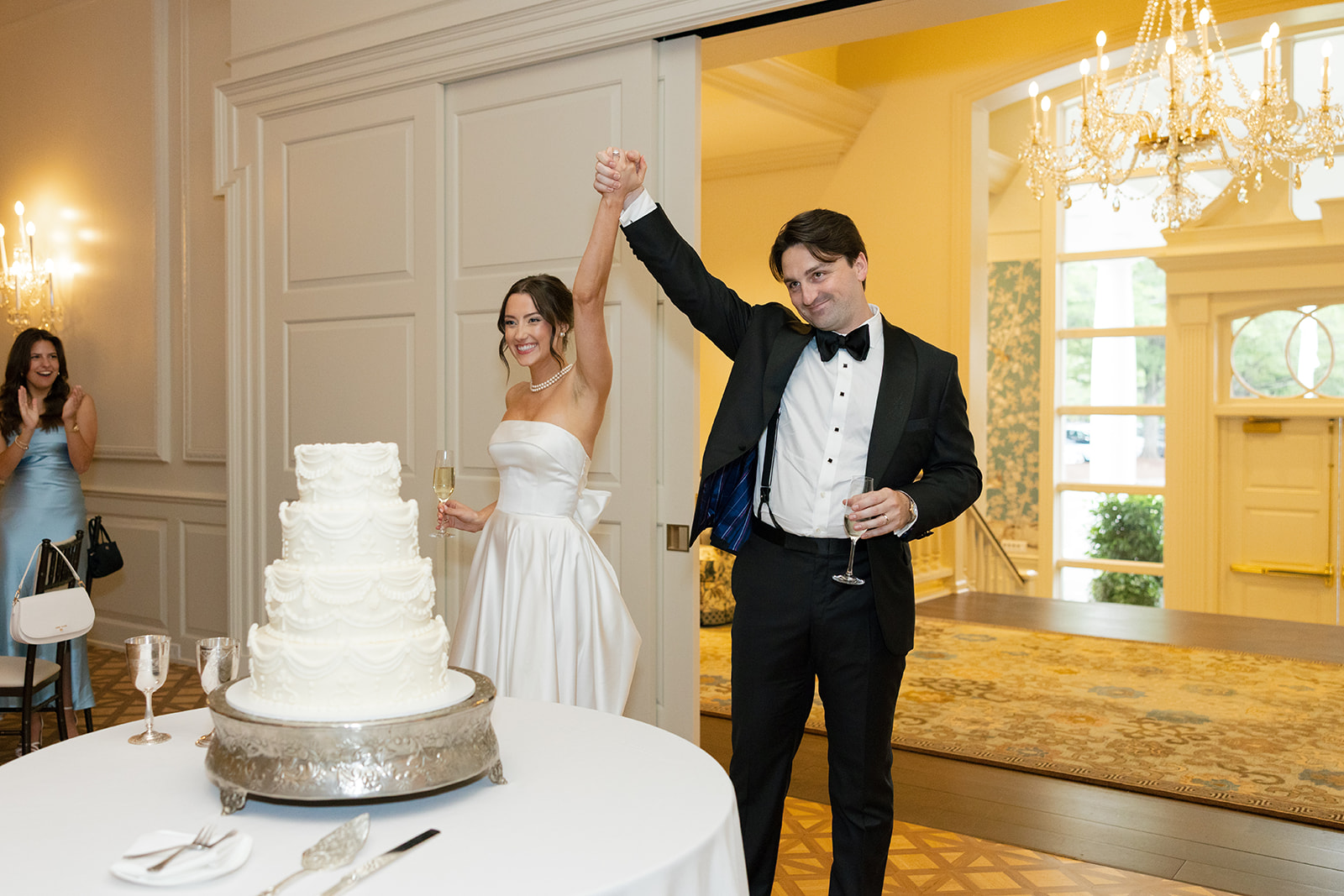 Bride and groom make a joyful entrance, raising their hands in celebration next to the wedding cake.